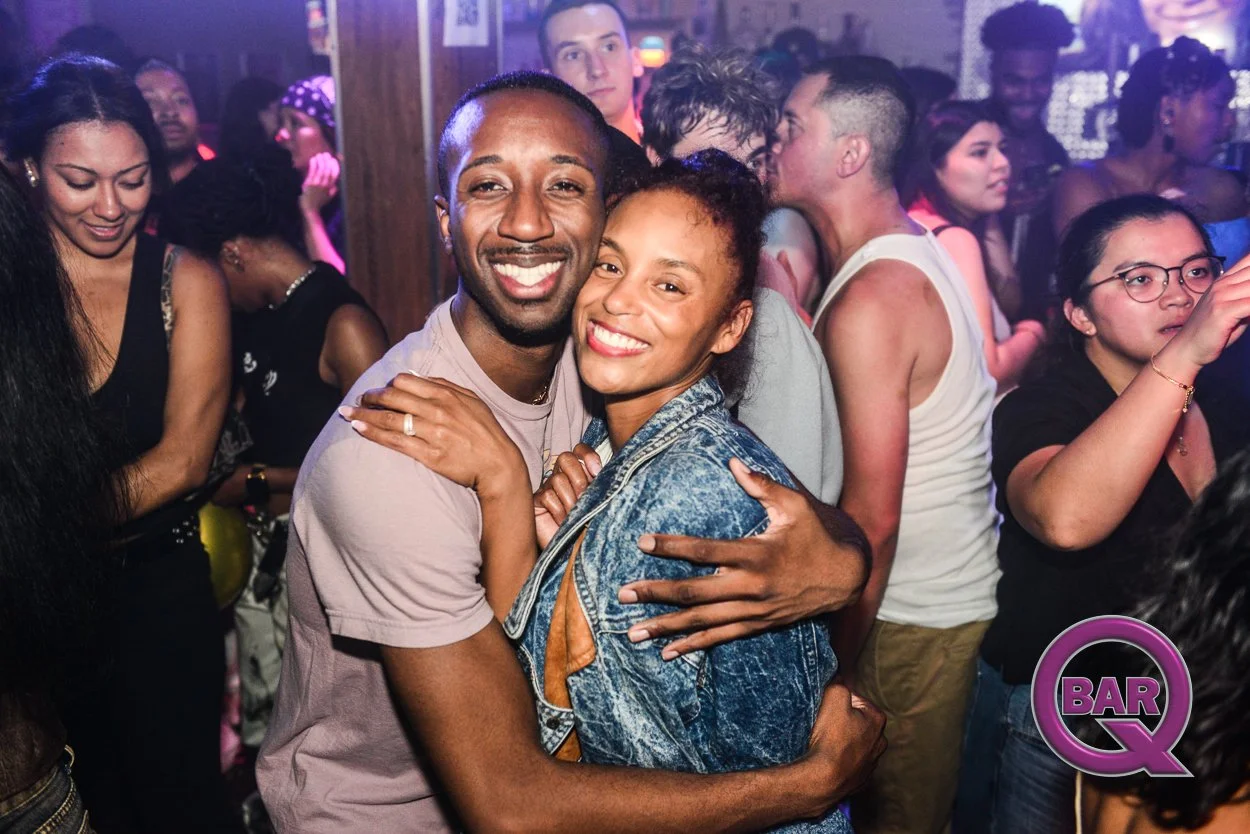 A joyful couple hugging and smiling at a lively indoor party or nightclub filled with diverse people dancing and enjoying themselves.