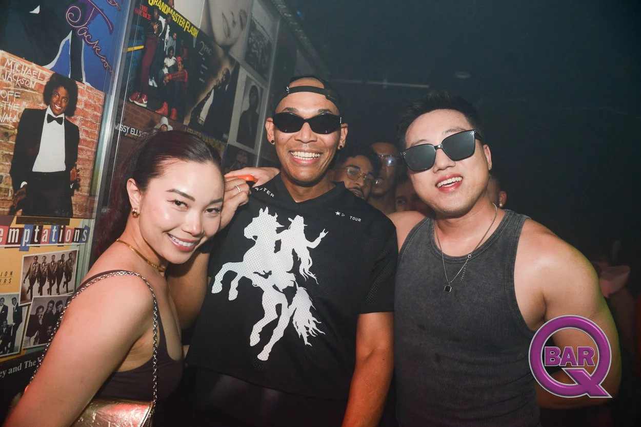 Three people smiling and wearing sunglasses at a bar, with posters on the wall behind them and a crowd in the background.