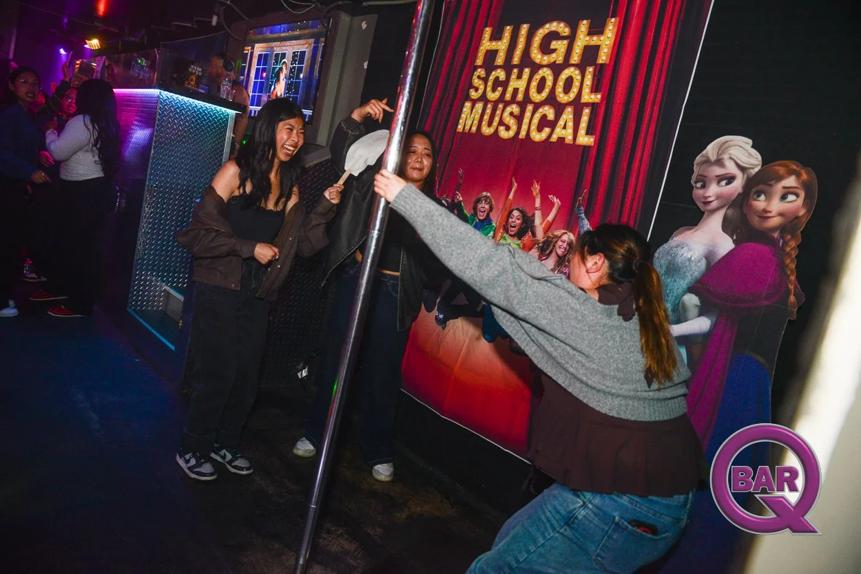 People dancing and laughing at a bar during a high school musical-themed event, with a large poster of Disney's Frozen characters Elsa and Anna on the wall.