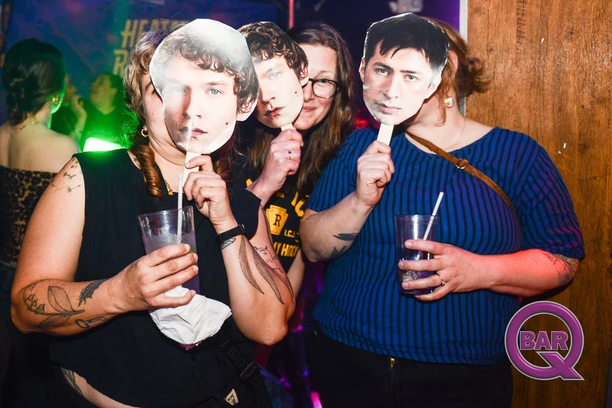 Four women at a bar wearing masks of male faces, holding drinks, and posing for the photo. The background is dimly lit with colorful nightclub lighting.