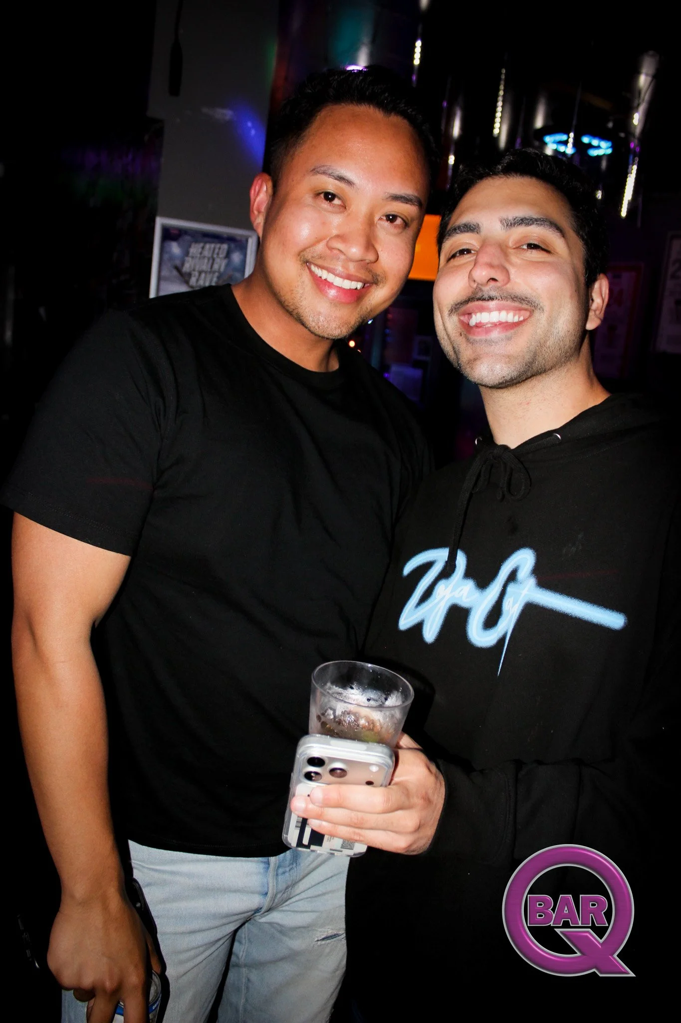 Two smiling men taking a selfie in a bar, one holding a drink and a phone, with bar signs and neon lights in the background.