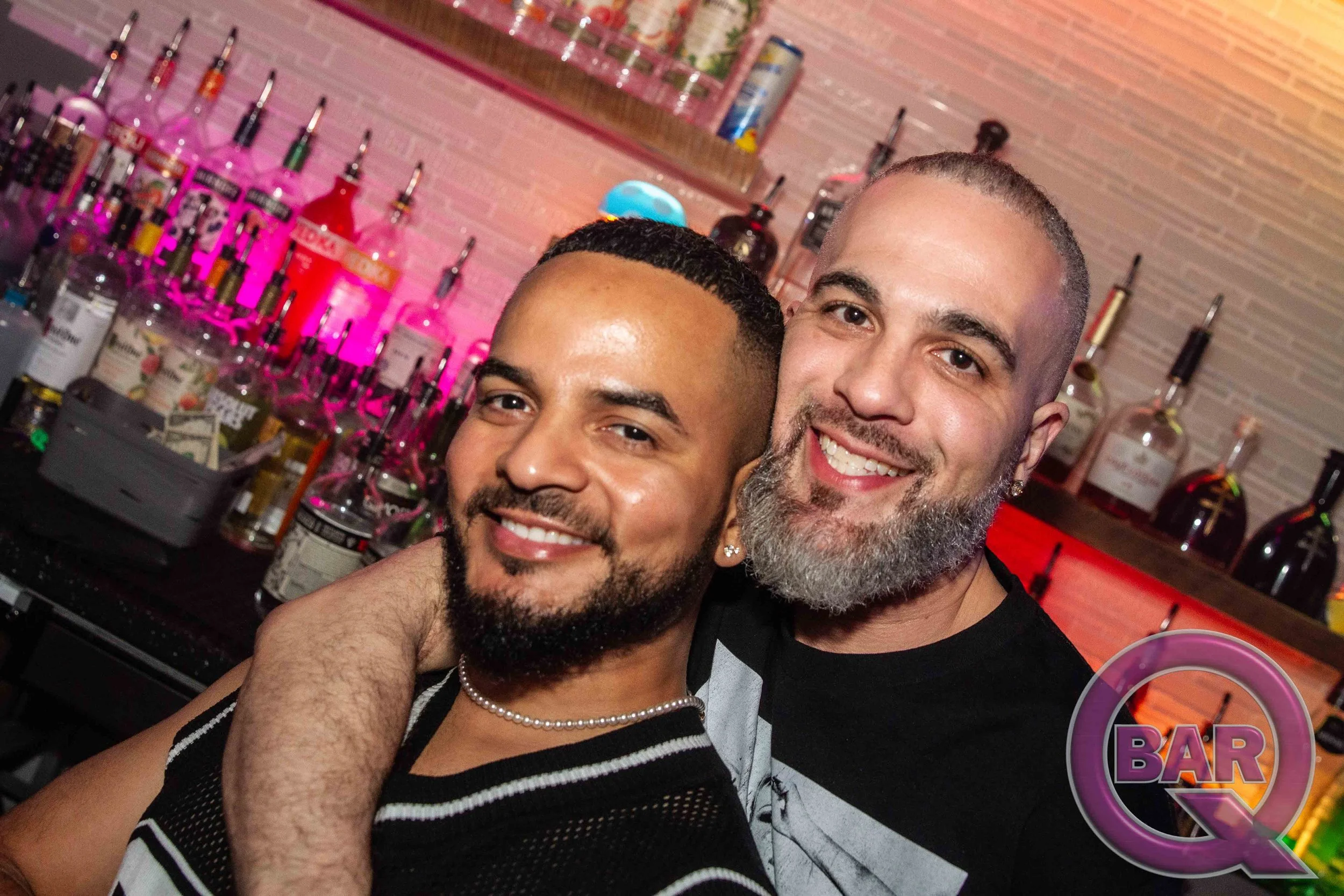Two men smiling, hugging in a bar with liquor bottles lined up on shelves behind them, pink and red lighting, one wearing a black and white striped shirt and pearl necklace, the other with a shaved head and beard.
