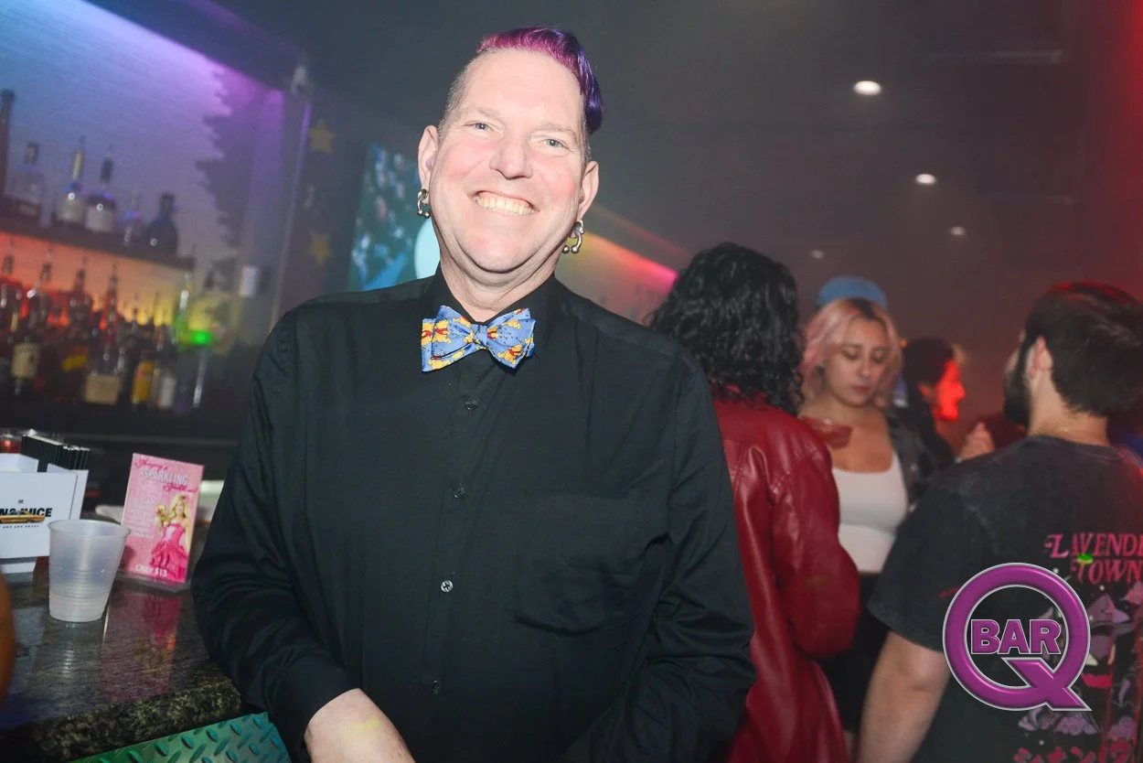 A smiling man with purple and green hair, wearing a black shirt and colorful bowtie, standing in a bar or nightclub with people in the background.