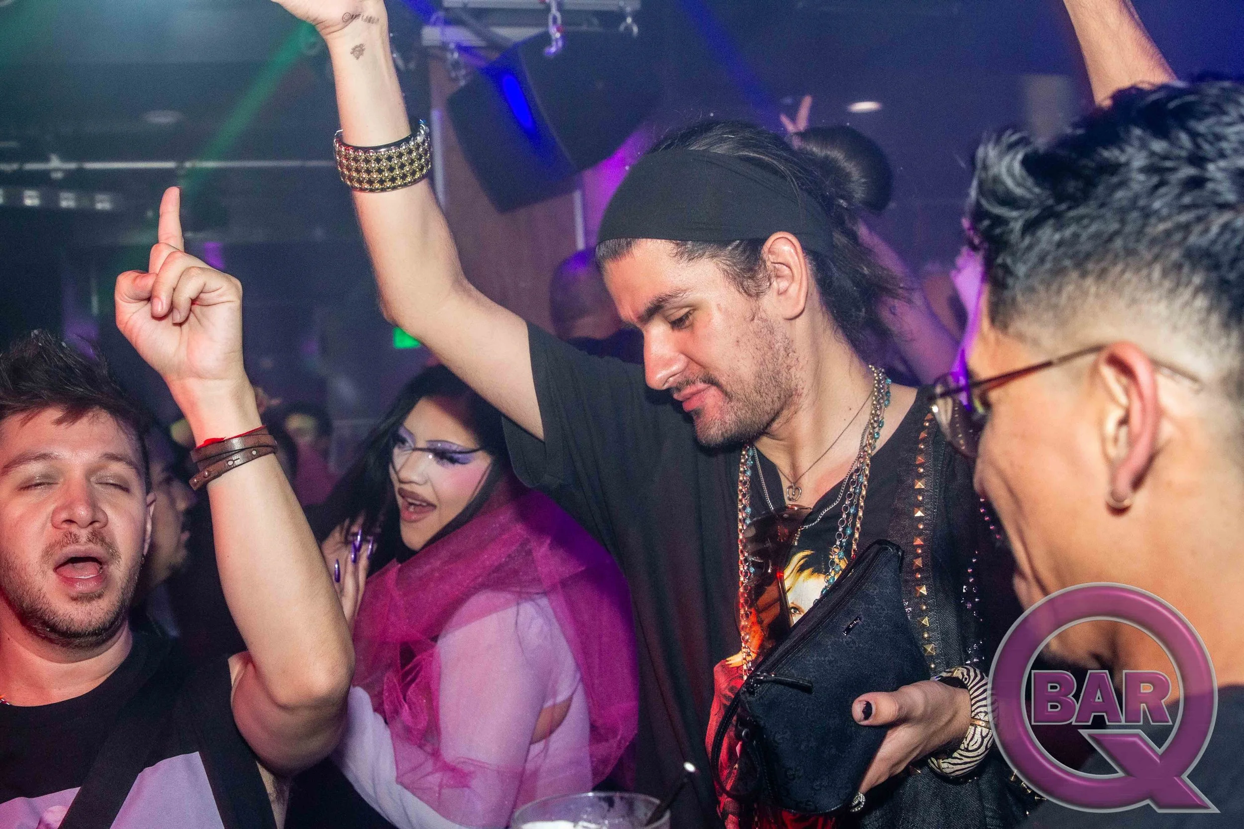 People dancing and enjoying at a nightclub or party with colorful lighting, including a man with long dark hair, a black headband, wearing a necklace and black clothing.