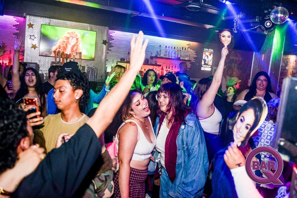 People dancing, singing, and socializing at a lively nightclub or bar with colorful lights, a large screen, and a bar counter in the background.