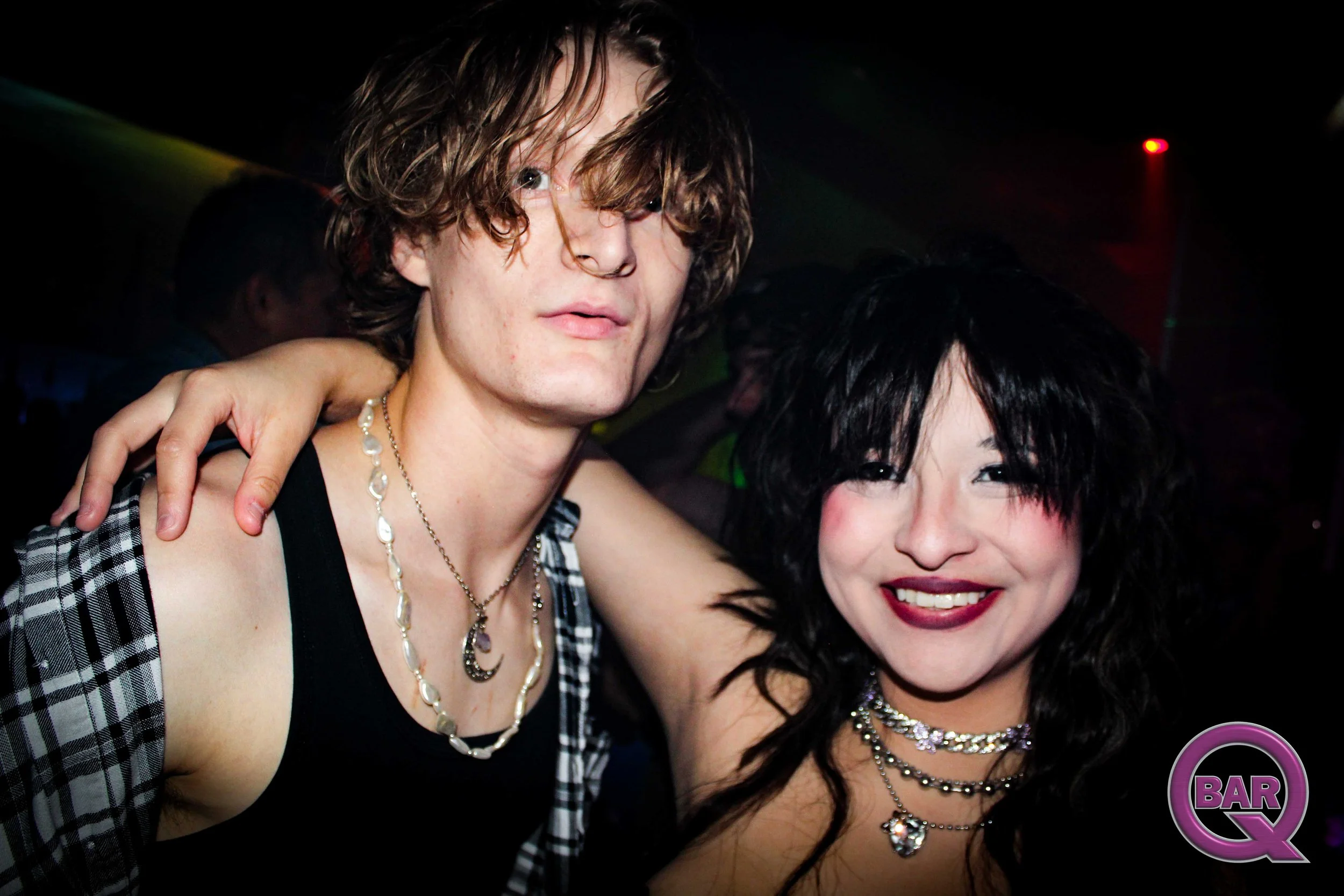 A young man and woman smiling and posing closely together in a dark environment with colorful lights, suggesting a nightclub or party setting.