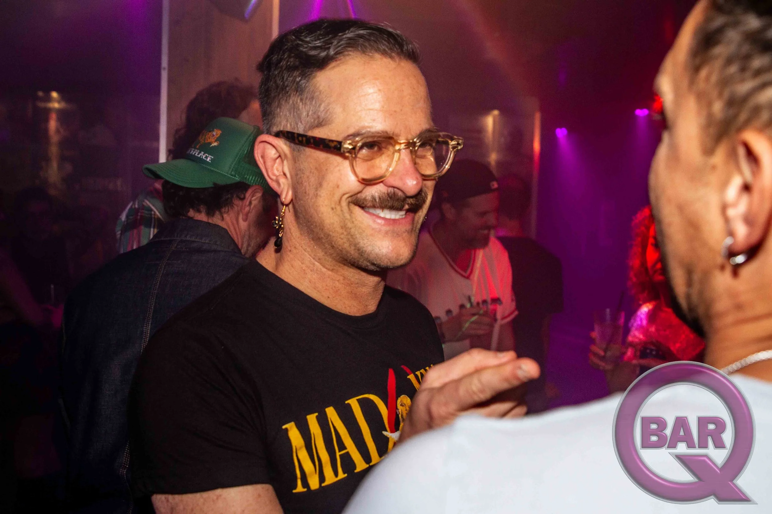 A man with glasses and earrings smiling at a bar, surrounded by people in a dimly lit venue with purple and red lighting.