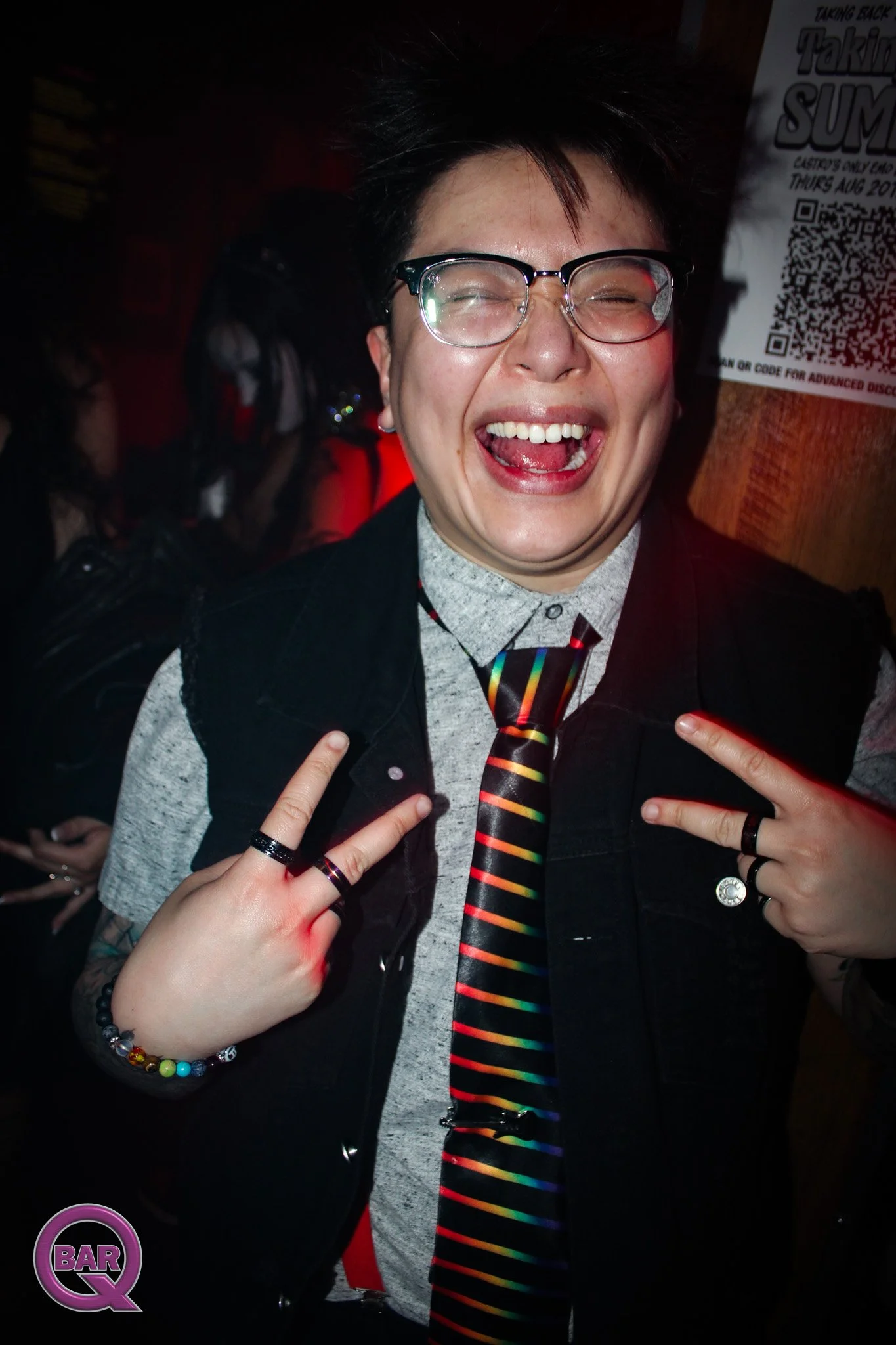 A person with short dark hair and glasses smiling widely, showing their tongue, with two hands making peace signs near their chest. The person is dressed in a shirt, a jacket, and has a rainbow-colored tie. Background is dark with a poster and other 