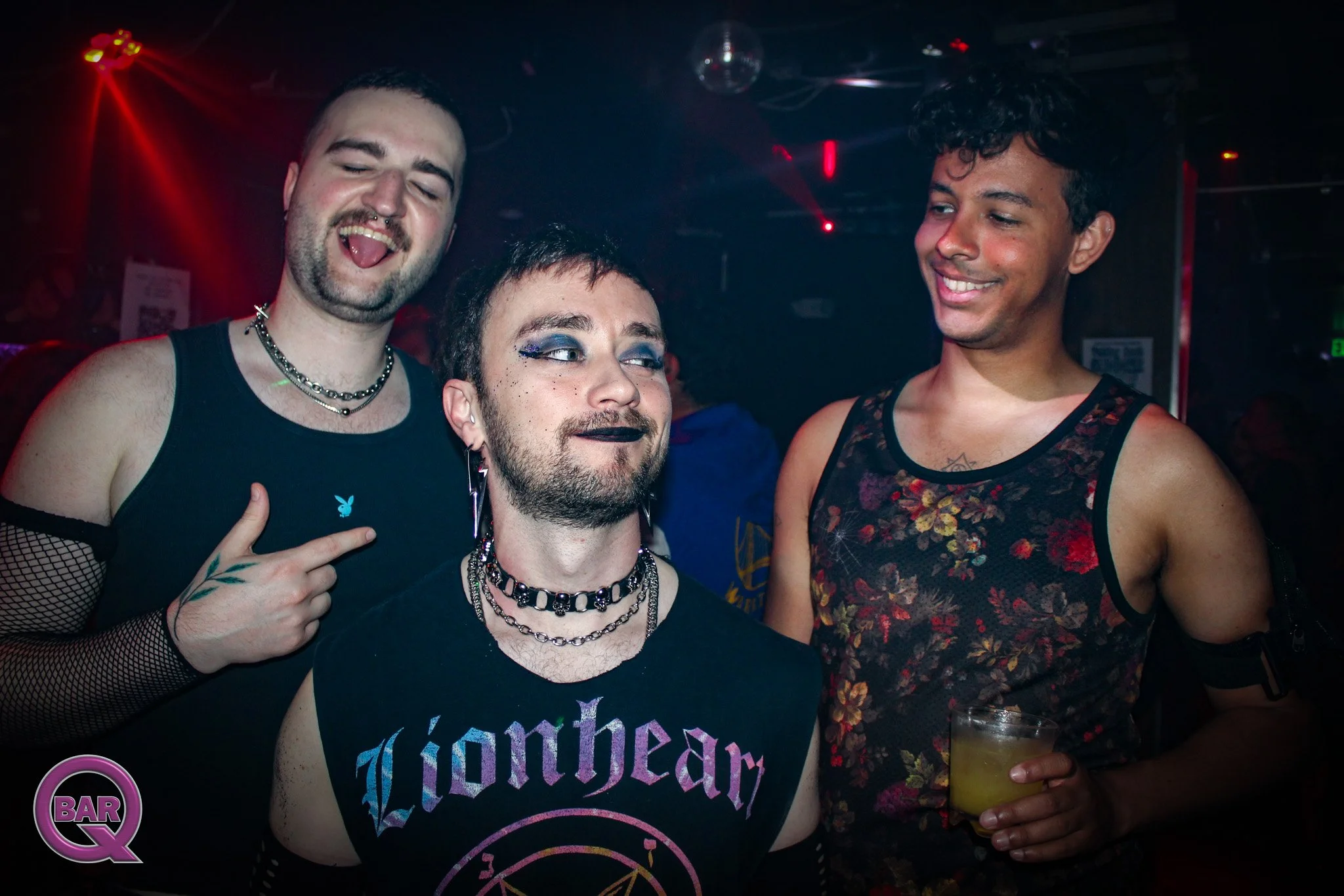 Three people at a nightclub, two men and one woman, all smiling. The man on the left has short dark hair, a beard, and is making a peace sign with his hand. The woman in the middle has makeup with glittery blue eyeshadow, black lipstick, and short da