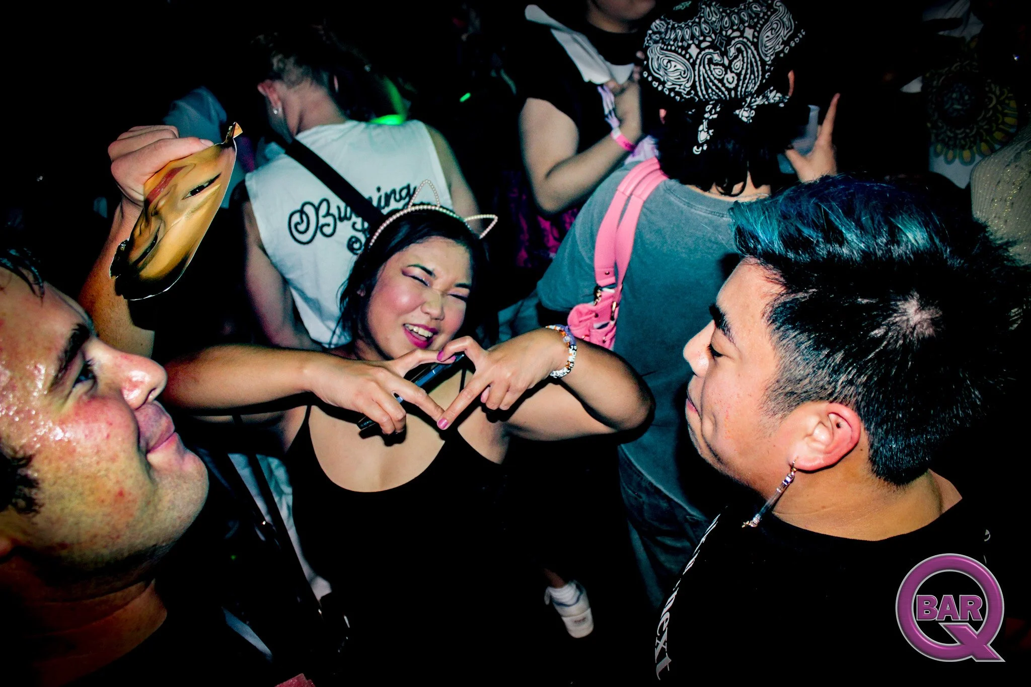 A group of people at a bar dancing closely together, some wearing accessories like a cat ear headband and a bandana, with one woman making a heart gesture with her hands in the center.