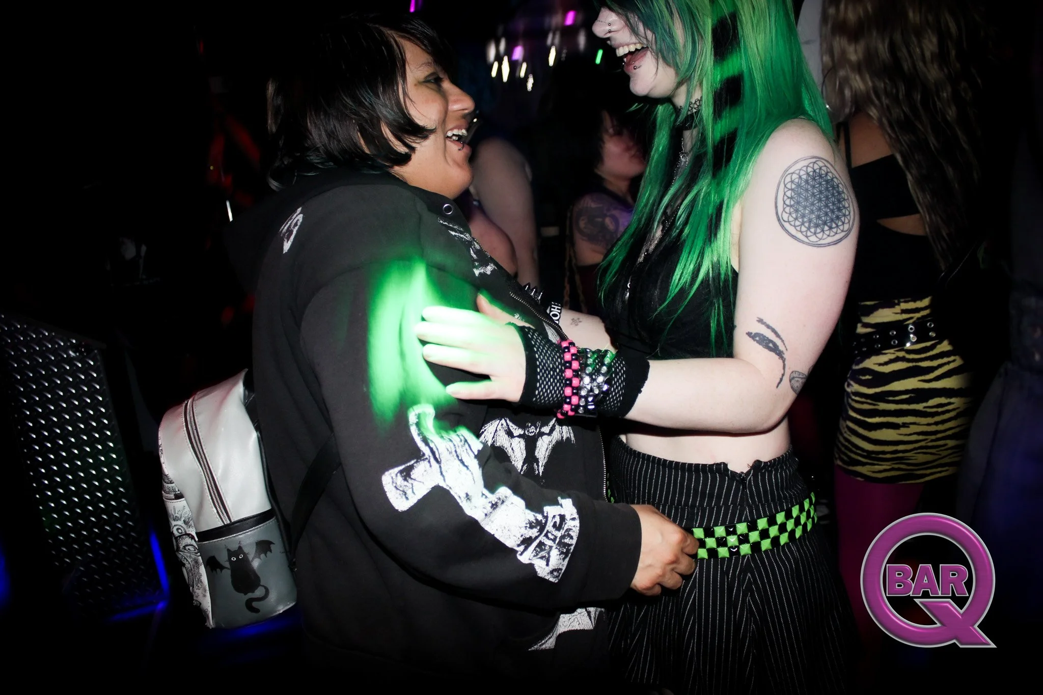 Two women are dancing and smiling at a nightclub, with one woman having dark hair and the other with bright green hair and tattoos, illuminated by colorful lights.