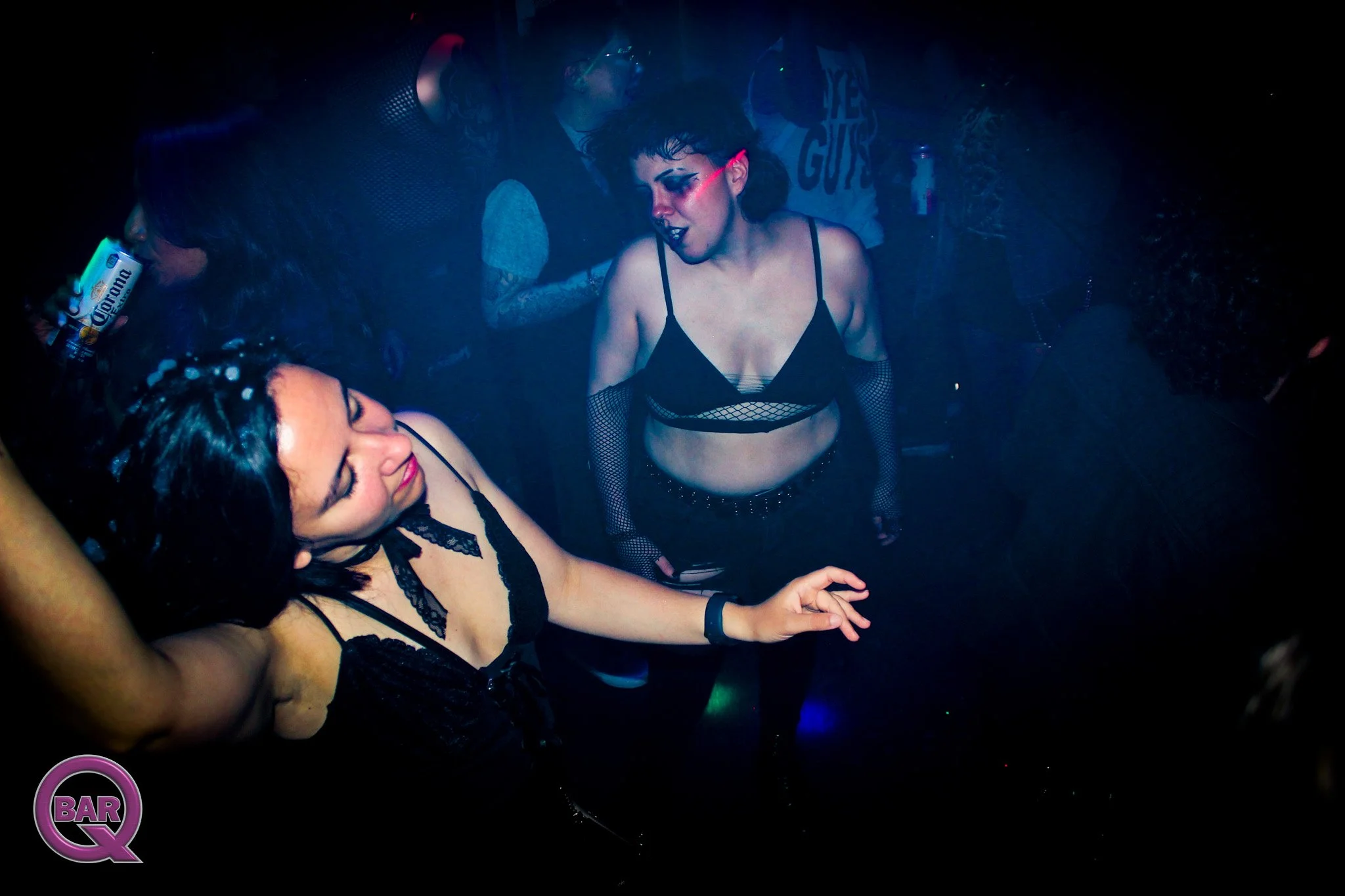 Women dancing at a club wearing revealing black outfits with fishnet details, illuminated by blue and purple lights.