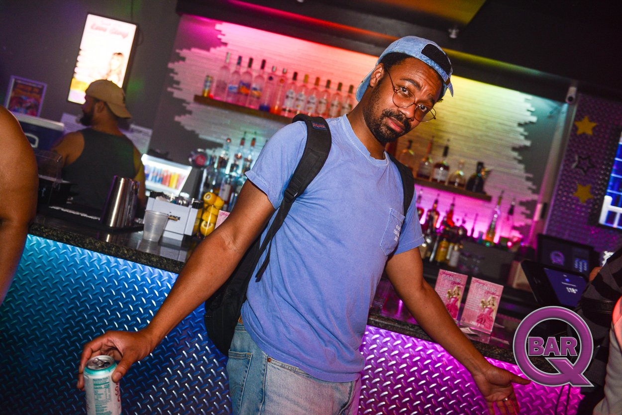 A man in a blue t-shirt and wearing glasses and a backwards cap stands at a bar, holding a soda can in his right hand. The bar has a colorful, neon-lit background with various bottles of alcohol on shelves.