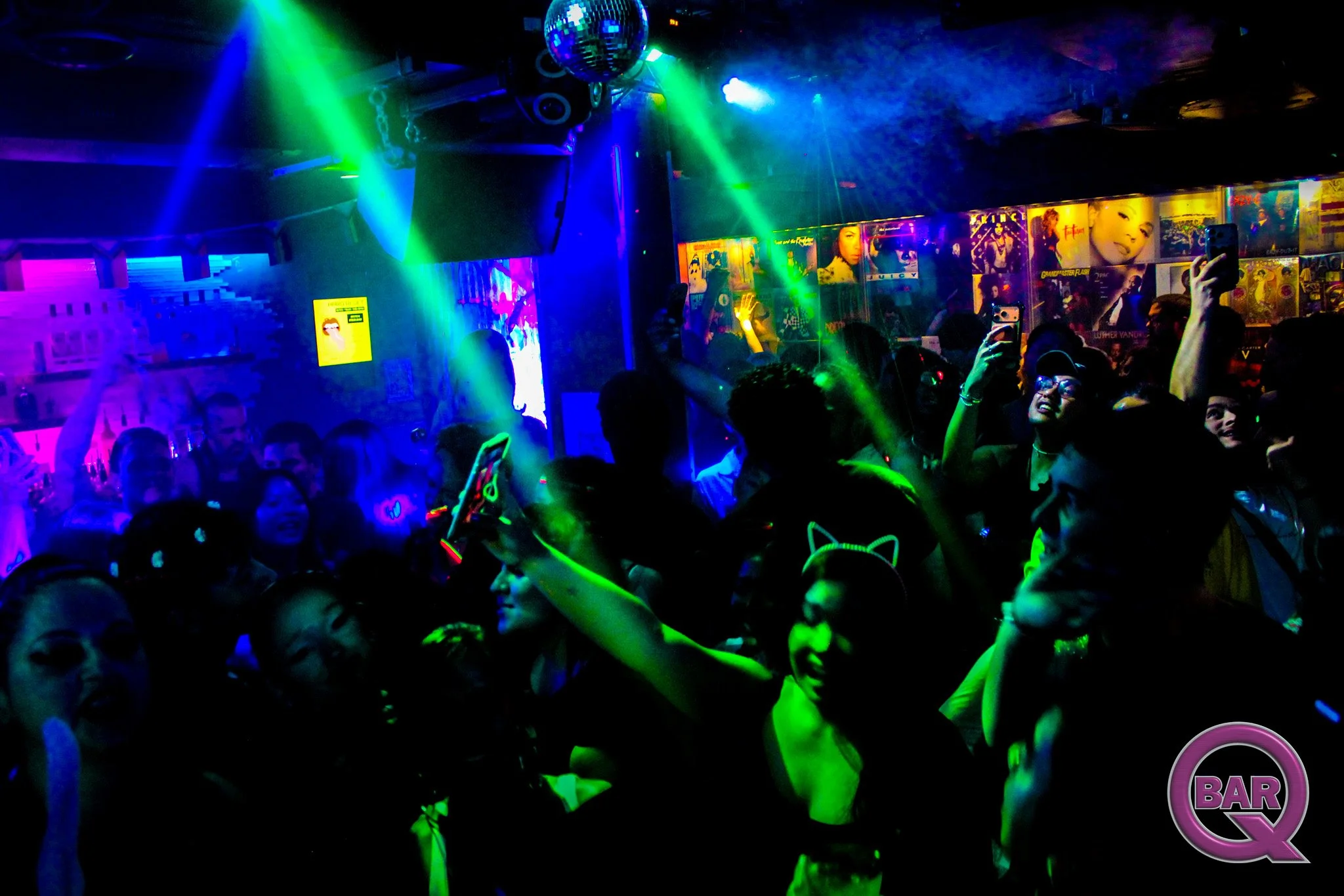 Crowd dancing and celebrating at a nightclub with colorful lights, a disco ball, and posters of famous women on the wall.