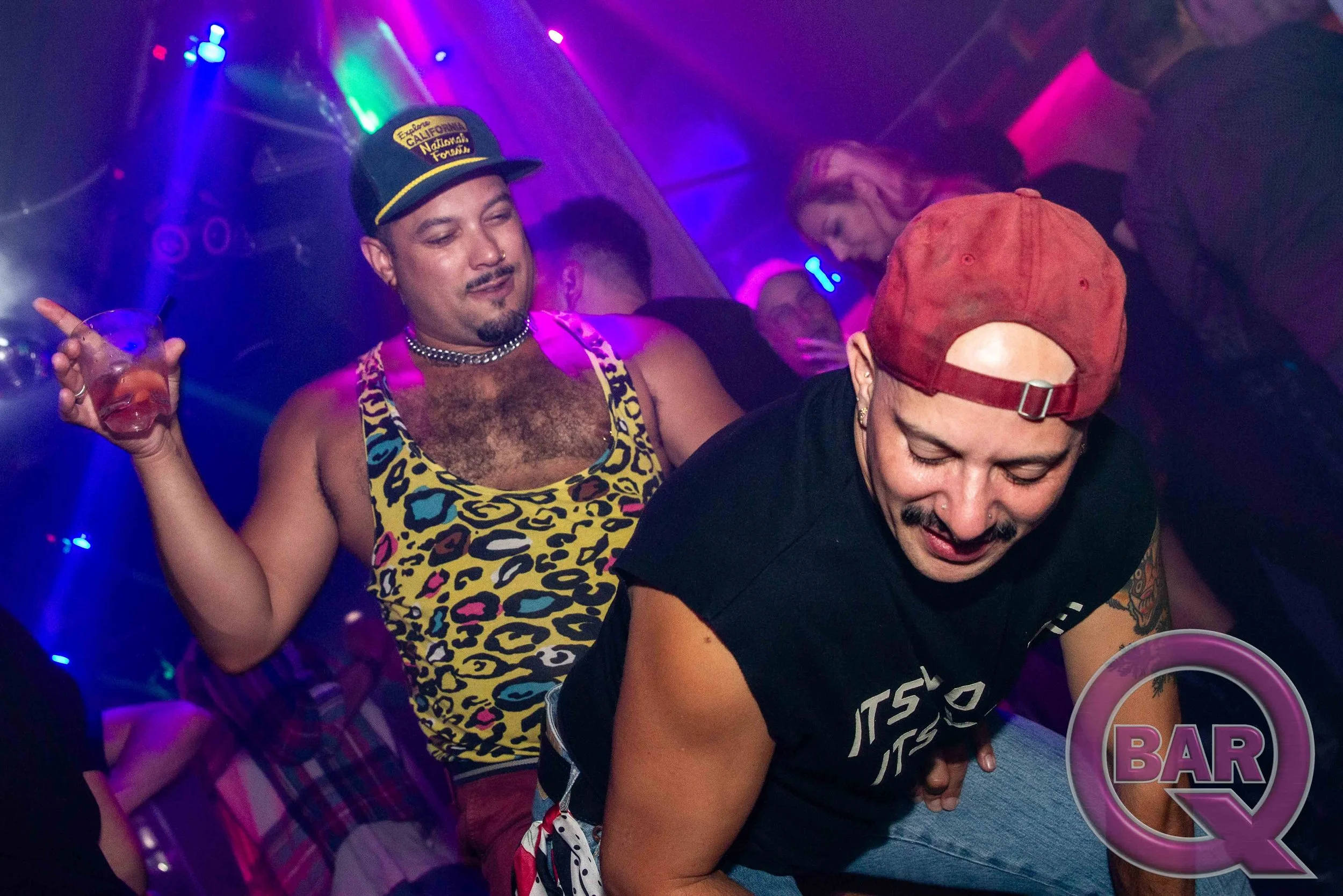 Two people dancing in a nightclub with colorful lights. One person is wearing a leopard print tank top, a black necklace, and a cap that says 'California National Forest.' The other person has a red backwards cap and is wearing a black shirt.