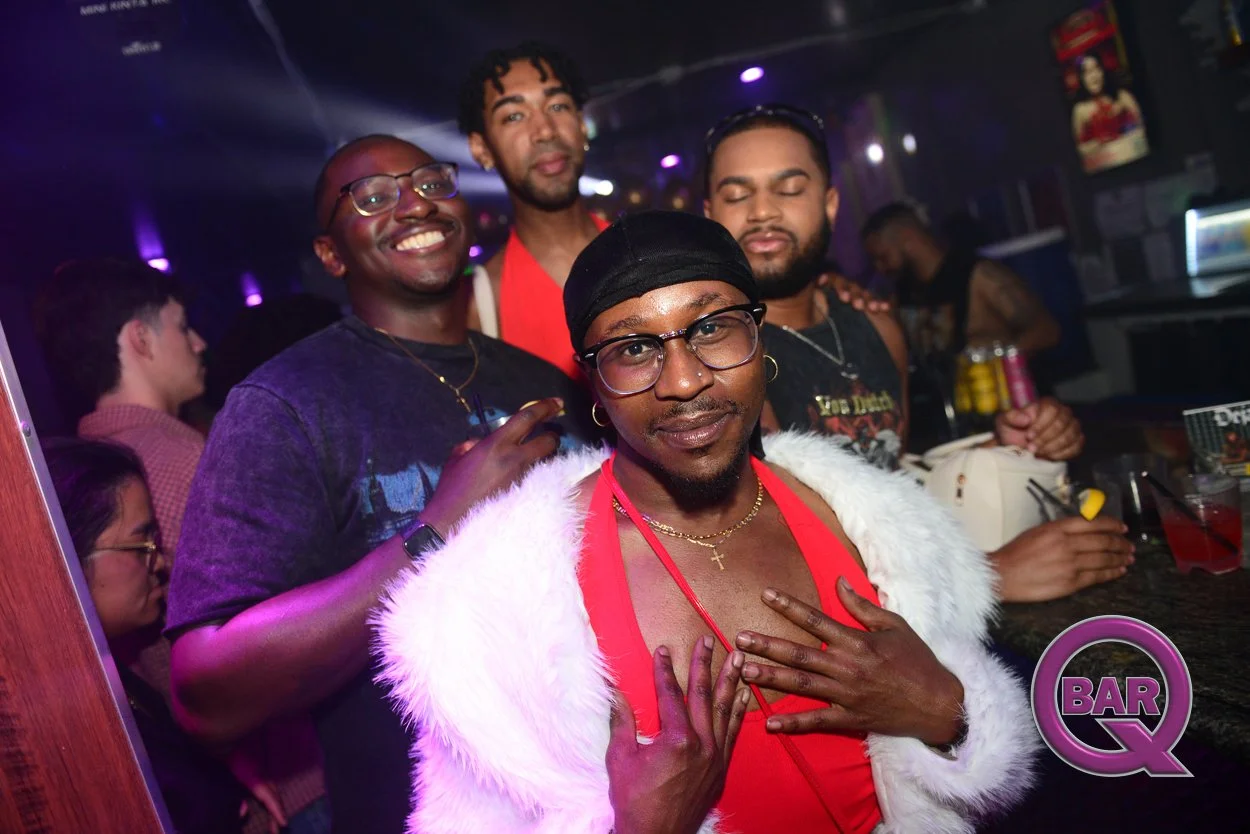 Four men posing in a bar with neon lighting, decorations, and drinks on the counter in the background. One man in the front wears a fur coat and a red shirt, with a hand on his chest. Others are smiling and standing behind him.
