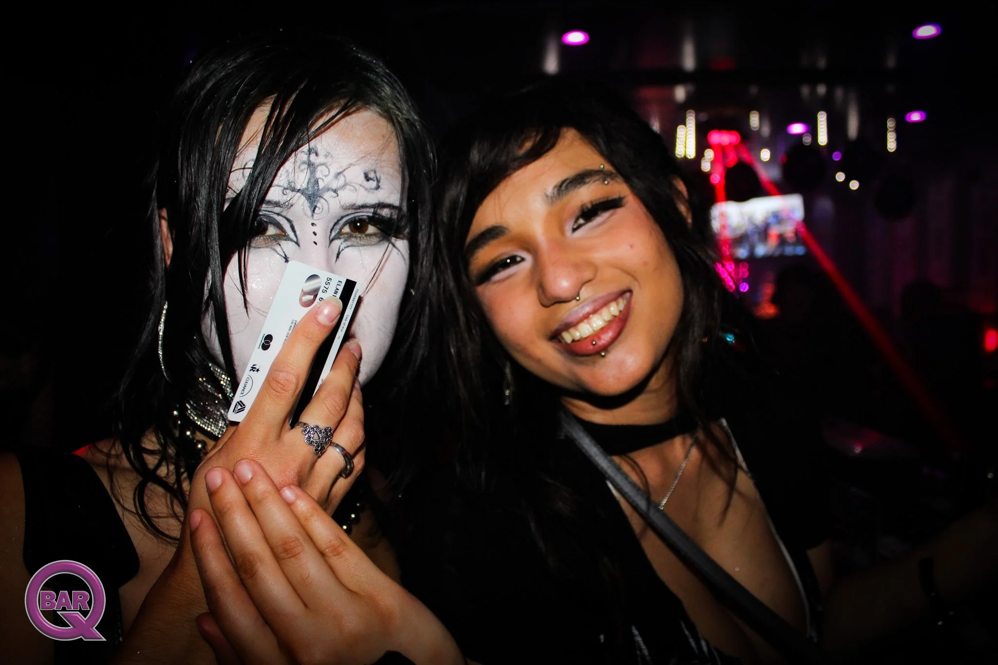 Two women smiling with hands together, one with face painted and holding a card, in a dark, neon-lit setting.
