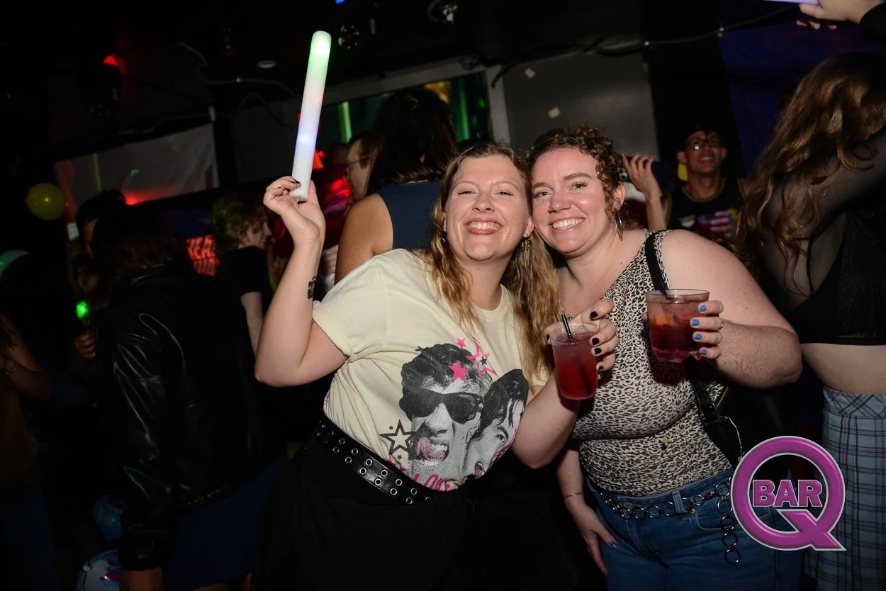Two women smiling and holding drinks at a party or bar with a crowd in the background, colorful lights, and a 'Bar Q' logo in the corner.
