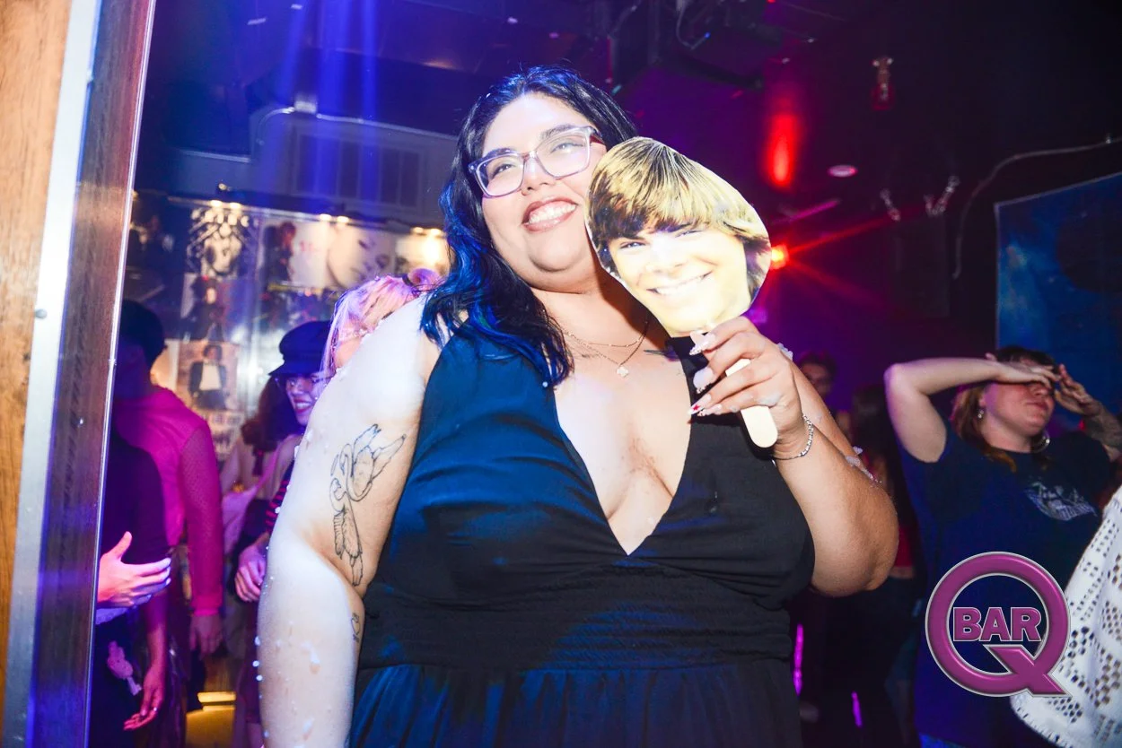 A woman with glasses and dark hair holding a paper mask with a smiling boy's face at a party or nightclub. The background shows other people dancing and having fun with colorful lights.