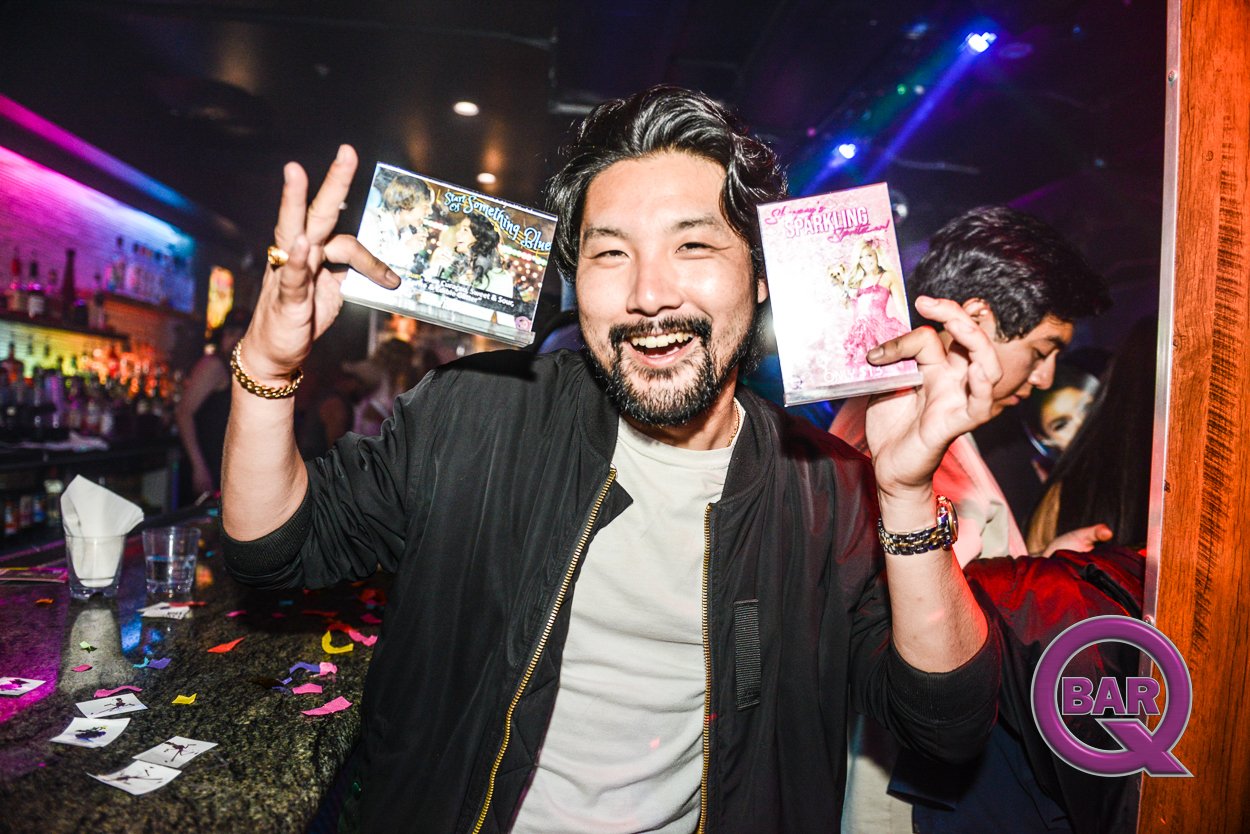 A man at a bar holding two DVDs, smiling and showing peace signs, with colorful party lighting and confetti on the counter.