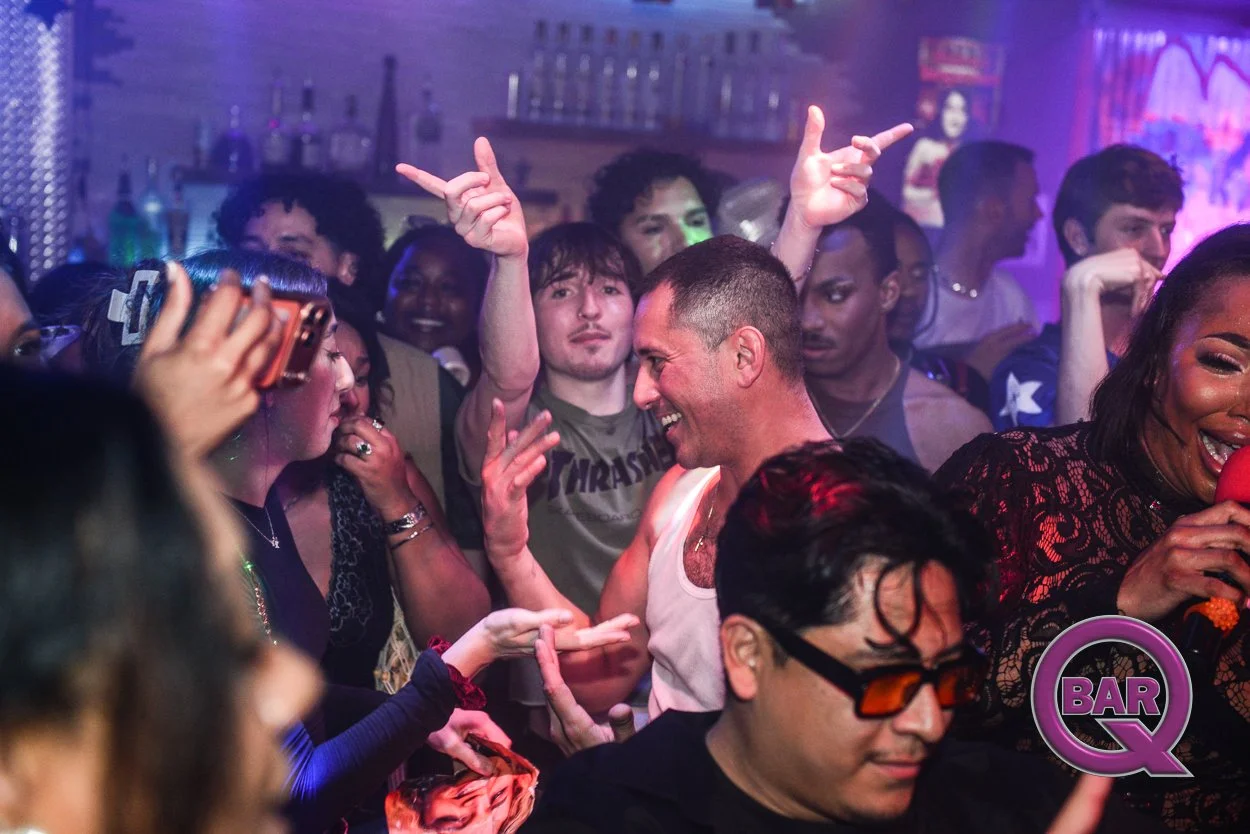 Crowd of people dancing and socializing in a nightclub with colorful lighting and a bar in the background.