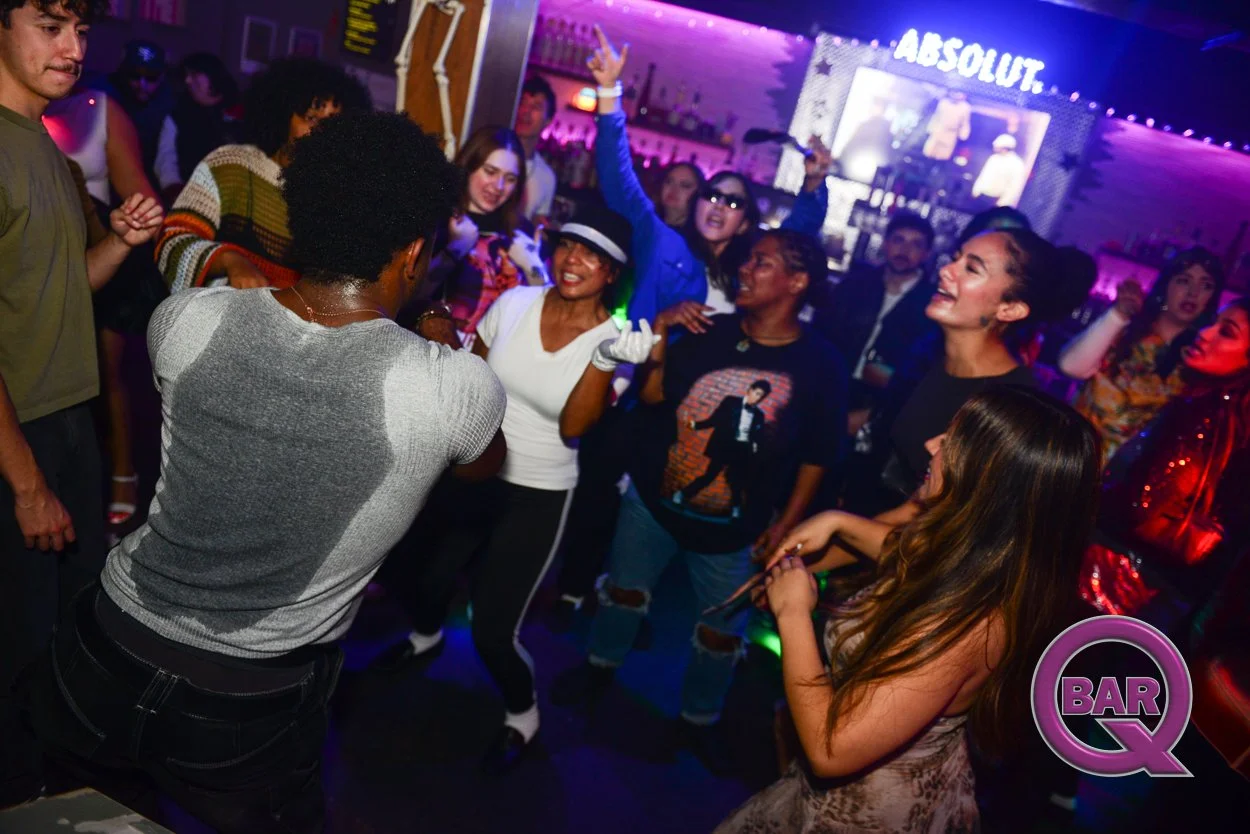 People dancing and having fun at a nightclub with colorful lights, a large screen, and a bar in the background.