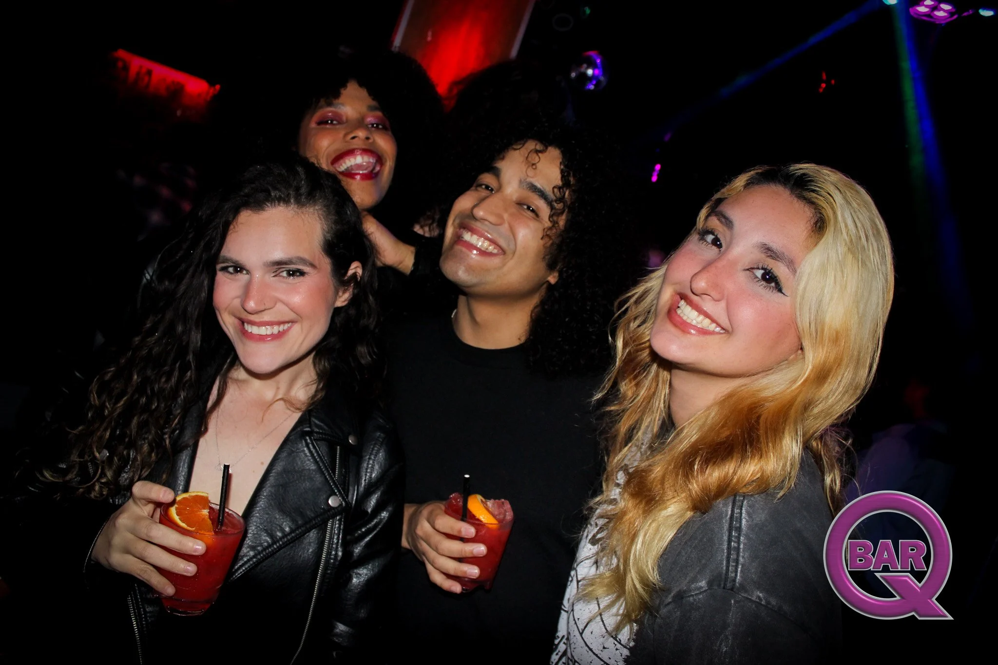 Four women smiling and celebrating in a nightclub holding drinks with orange slices, against a dark background with colorful lights.