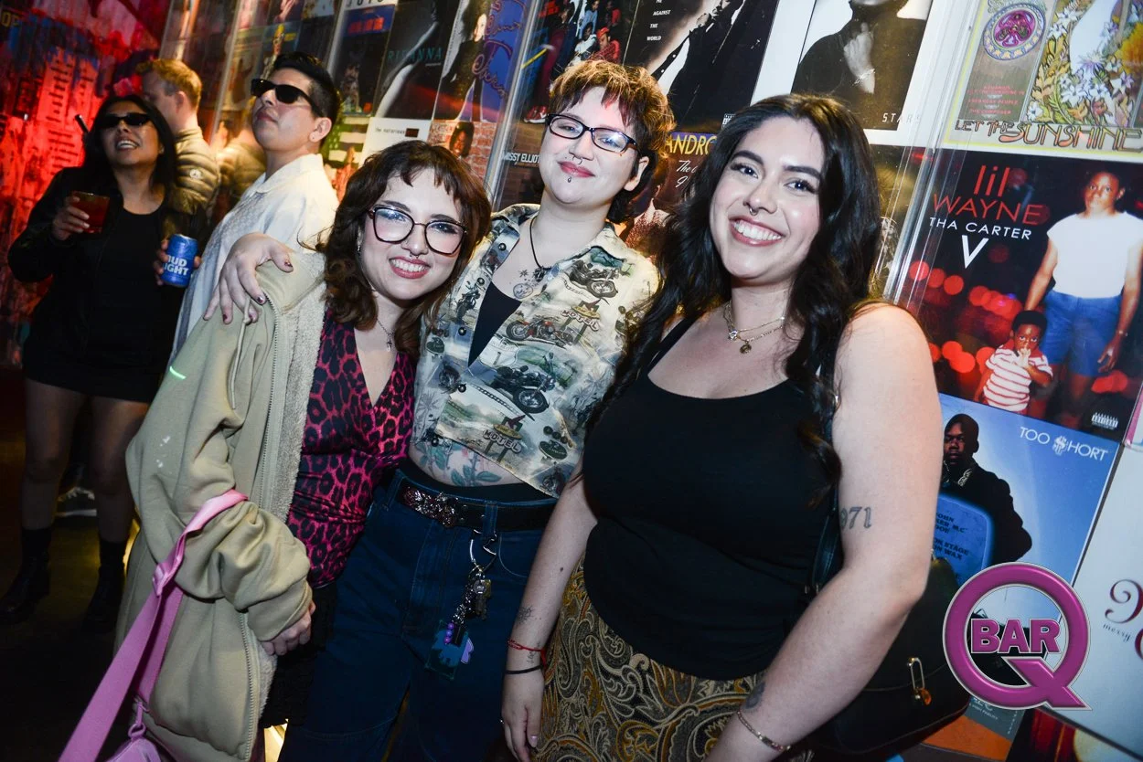 A group of five people posing together in front of a wall decorated with posters at a music or entertainment venue. Four women and one man in the background are also visible, holding drinks and wearing casual attire with some wearing sunglasses.