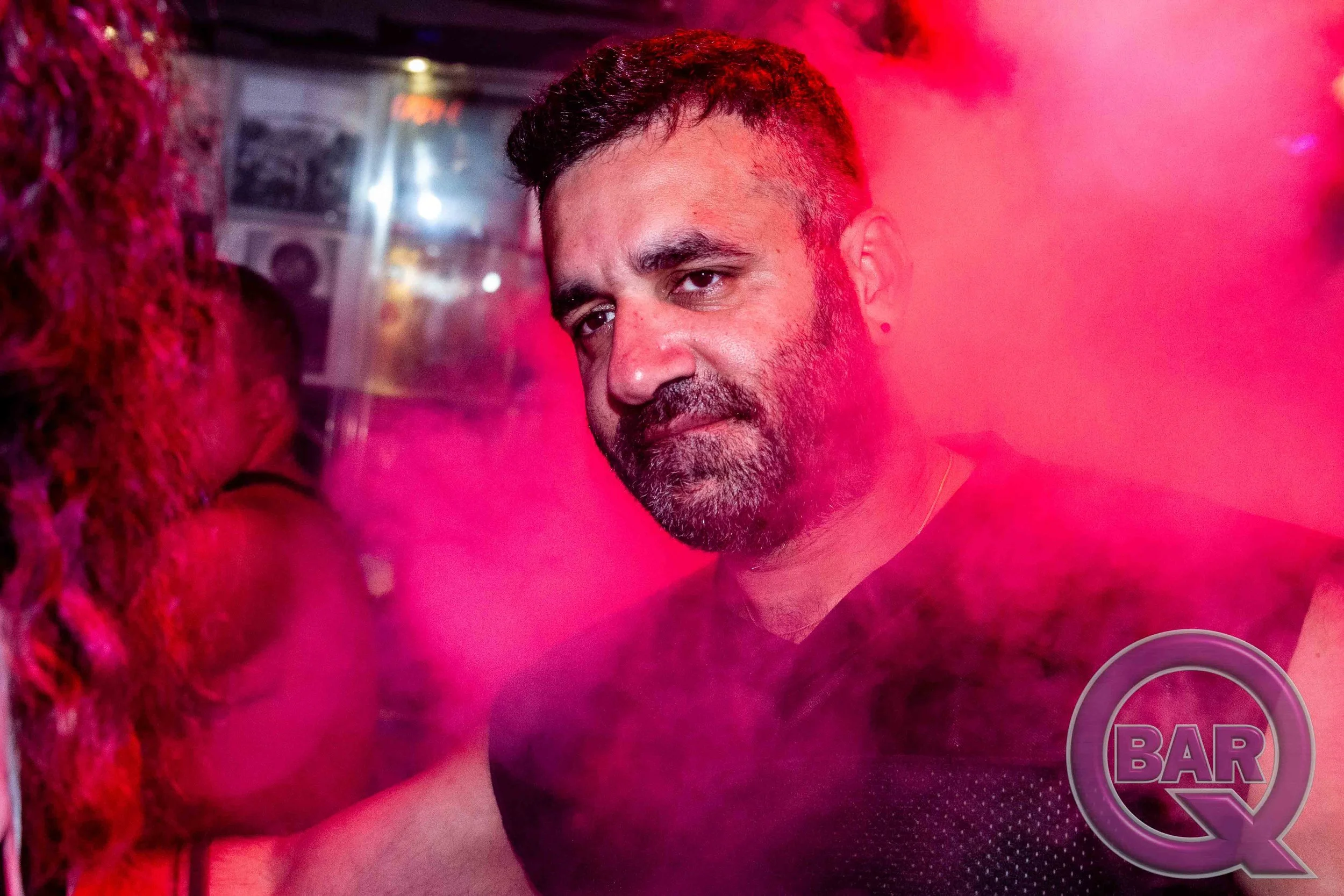 A man with short dark hair and a beard looking at the camera, surrounded by pink fog or smoke in a nightclub or bar setting.