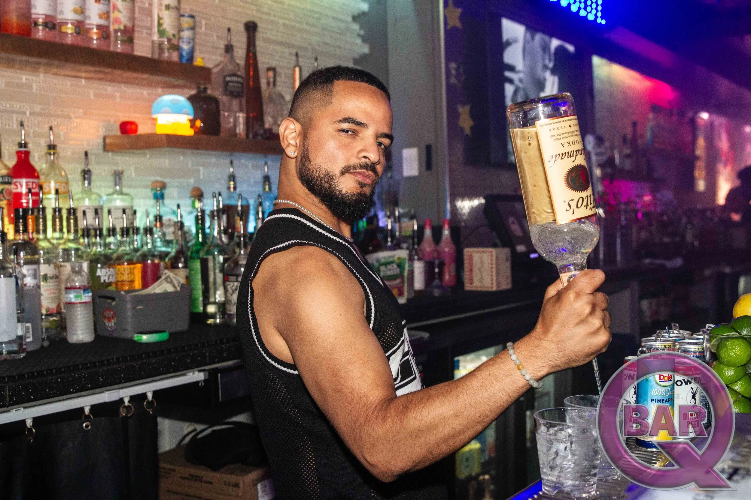 A man with a beard and short hair, wearing a striped sleeveless shirt, holding a wine glass, standing behind a bar with various bottles and mixers, in a dimly lit setting with colorful neon lights.