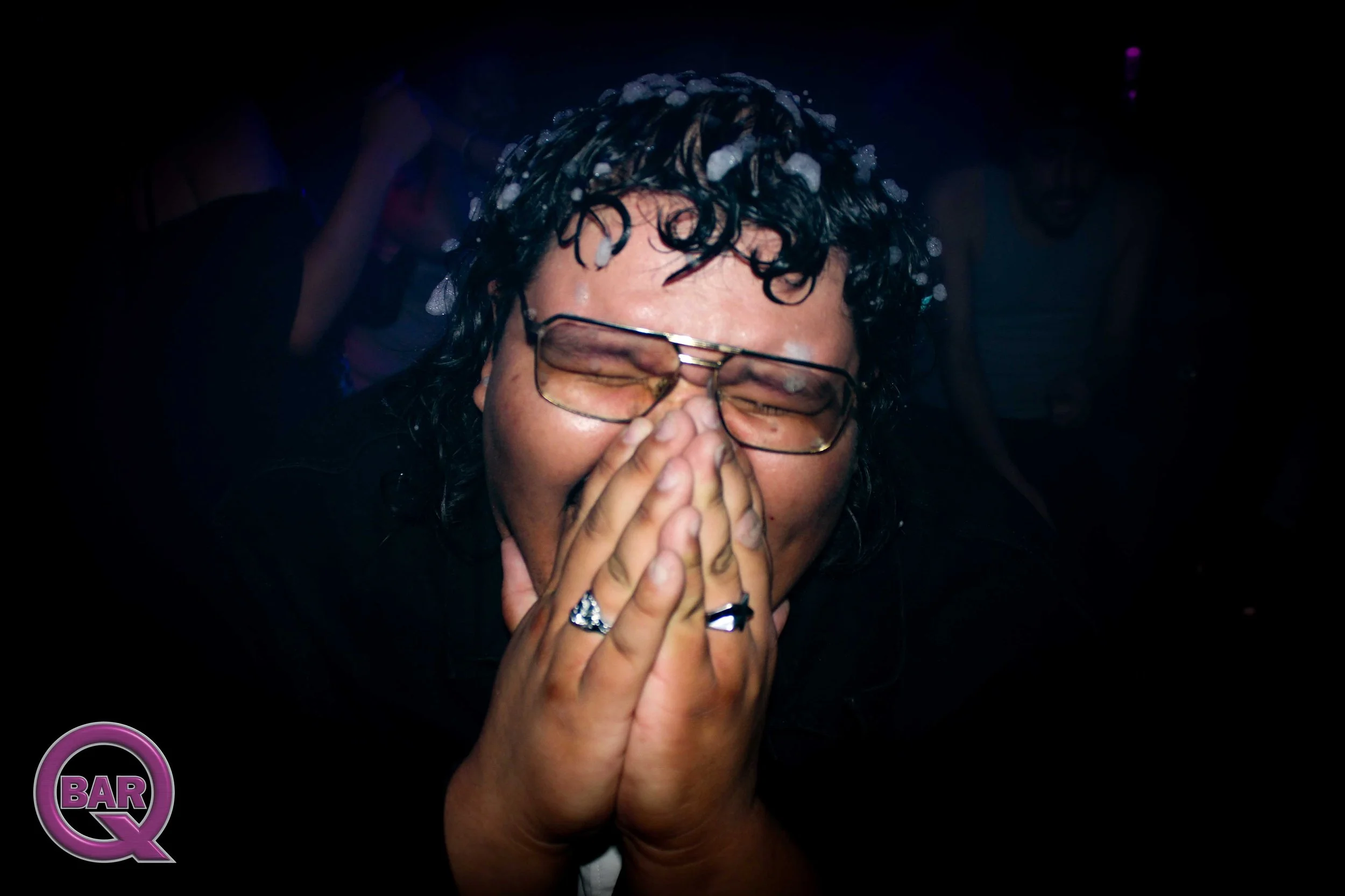A person with black, curly hair is smiling with eyes closed, hands covering their face, wearing glasses and rings, in a dark setting with people in the background, illuminated by colorful lighting.