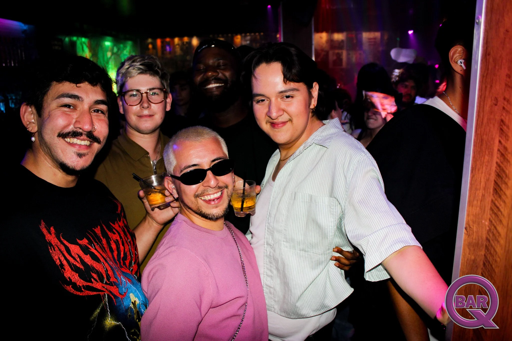 Group of people at a bar, smiling and holding drinks in a dimly lit nightclub with colorful lighting.