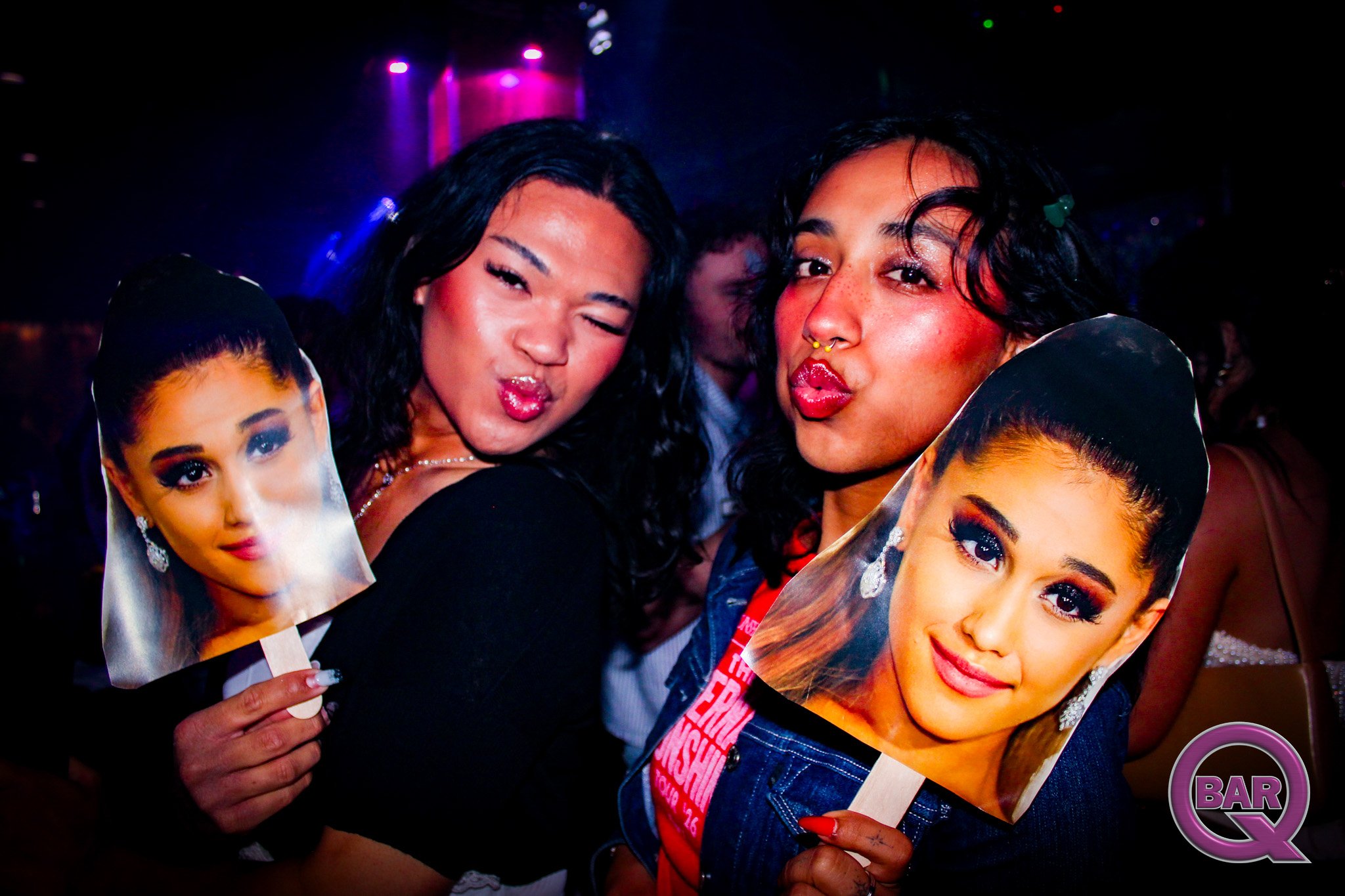 Two women at a nightclub holding printed photos of a woman with makeup and earrings, with colorful lights in the background.