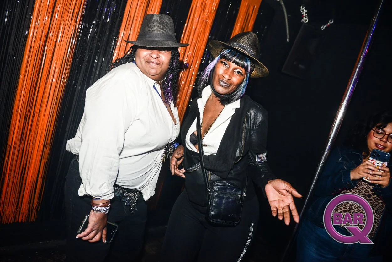 Two women wearing black hats and black outfits posing inside a nightclub with wooden wall decor. One woman has purple hair and is making a gesture with her hand, while another woman in the background is taking a photo with her phone.
