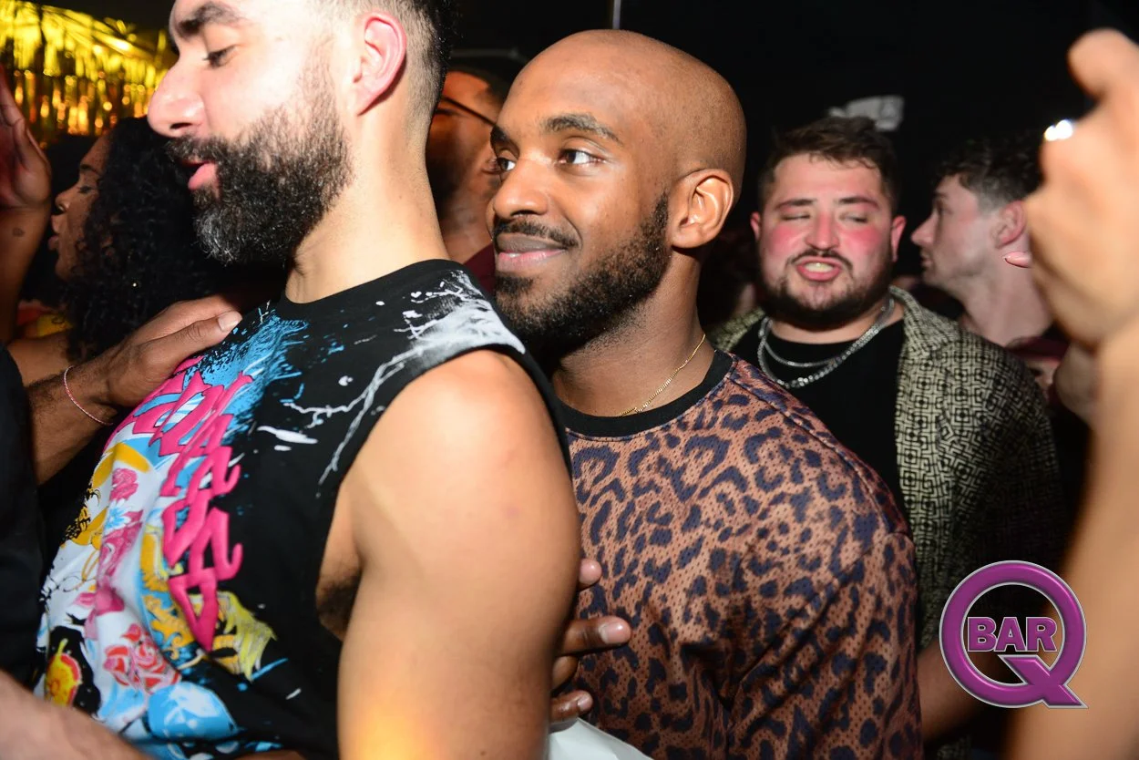 People dancing and socializing at a nightclub or bar, with a man in a leopard print shirt and a man with a beard and a vivid colorful shirt in the foreground, and others in the background.