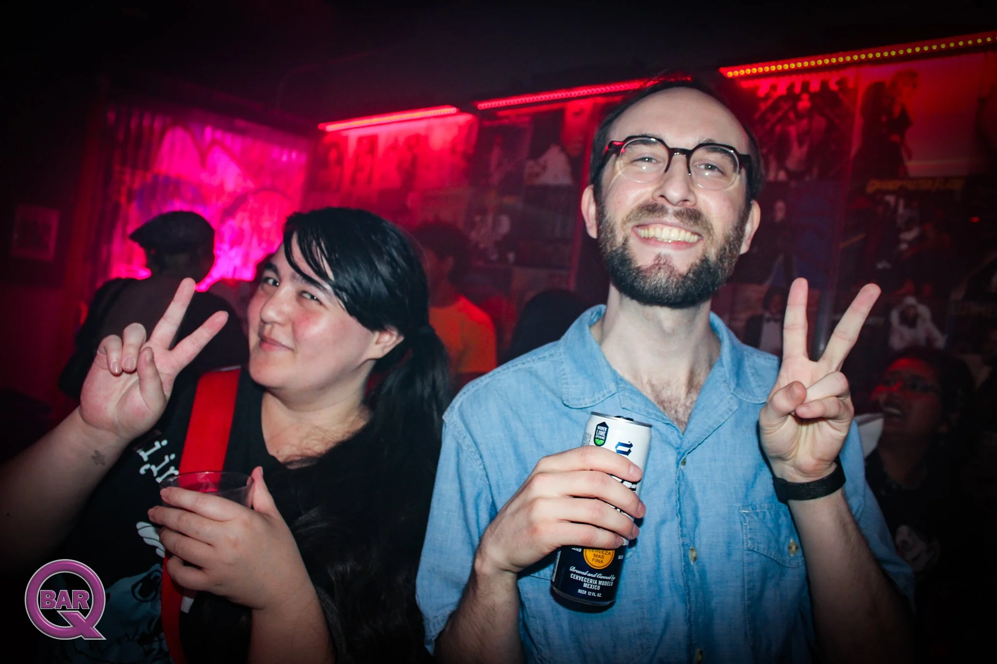 Two smiling people at a bar or party, making peace signs with their hands. The man has glasses and a beard, holding a beer can. The woman has dark hair and is wearing a black shirt with red accents. The background has dark lighting and colorful neon 