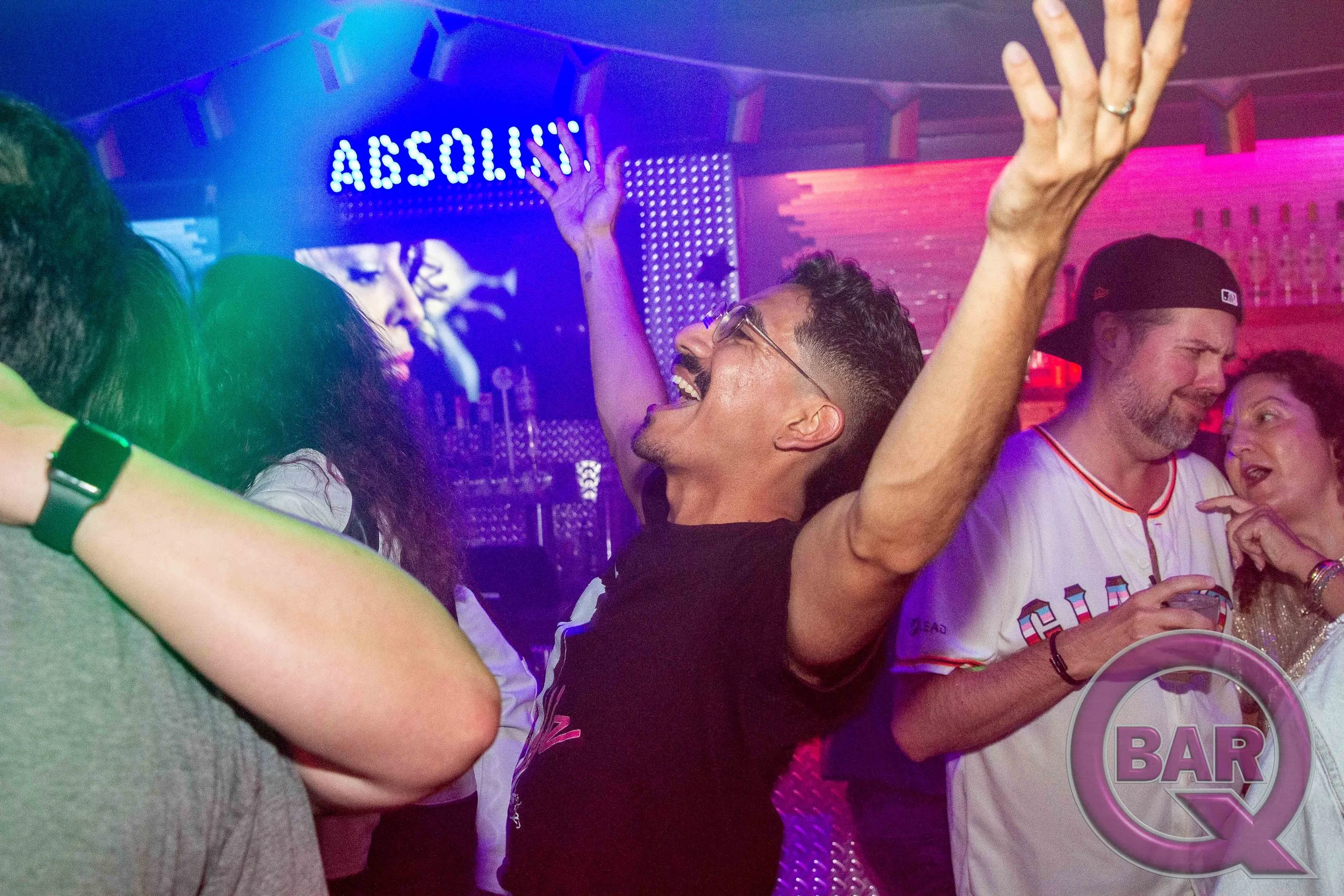 People dancing and socializing at a bar with neon and colorful lighting, with a sign that says 'ABSOLUT' in the background.
