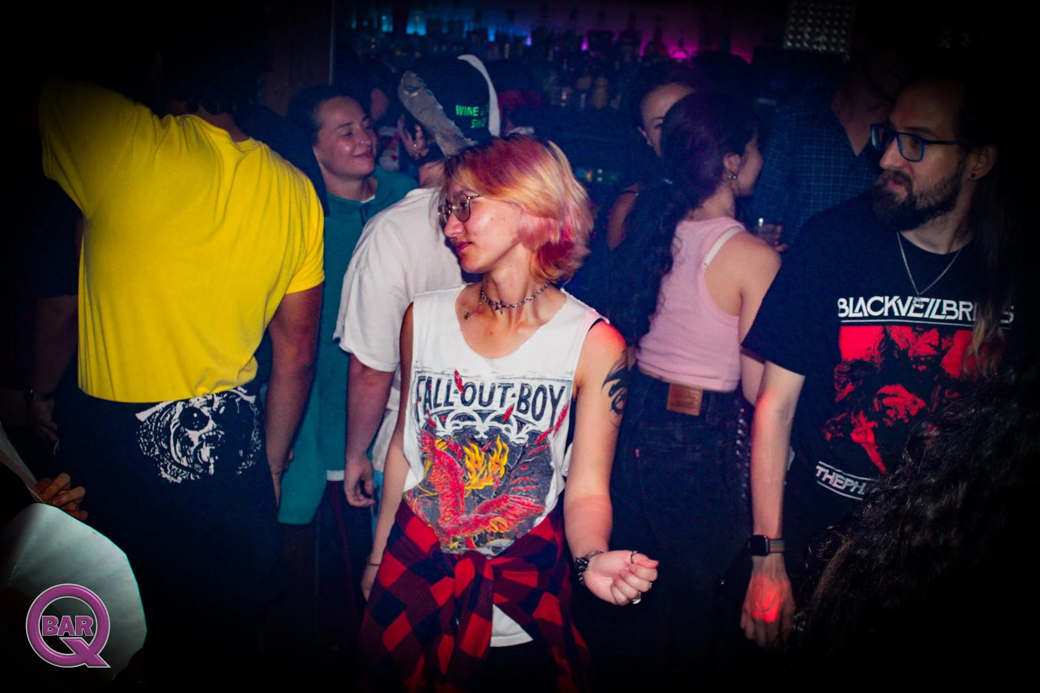 People dancing and socializing at a nightclub with colorful lighting, a bar sign, and a DJ booth in the background.