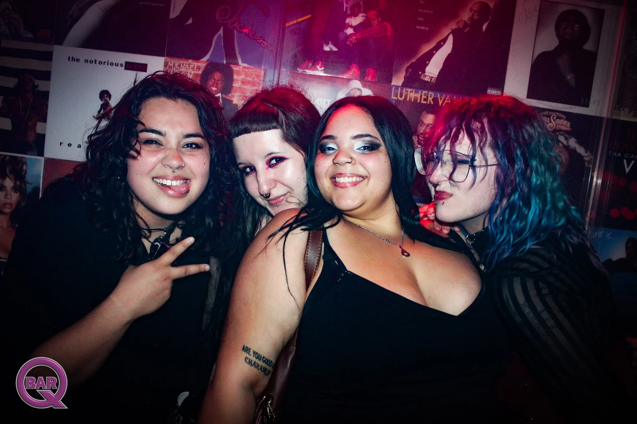 Four women pose for a photo inside a bar or club. They have dark hair, pierced noses, and are smiling or making playful gestures. The background features posters of musicians and a purple and black themed decor.