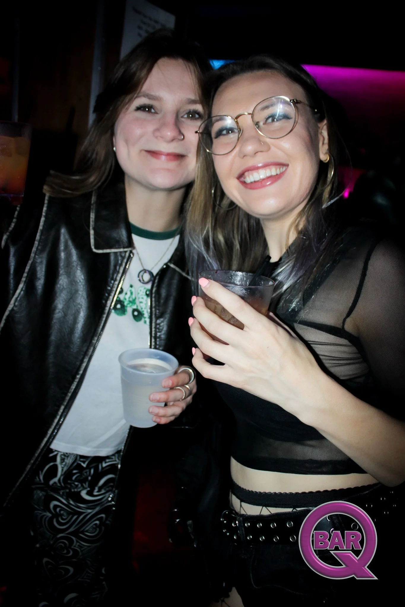 Two women smiling and posing for a photo at a bar or nightclub, holding drinks in their hands, with tinted glasses and stylish clothing.