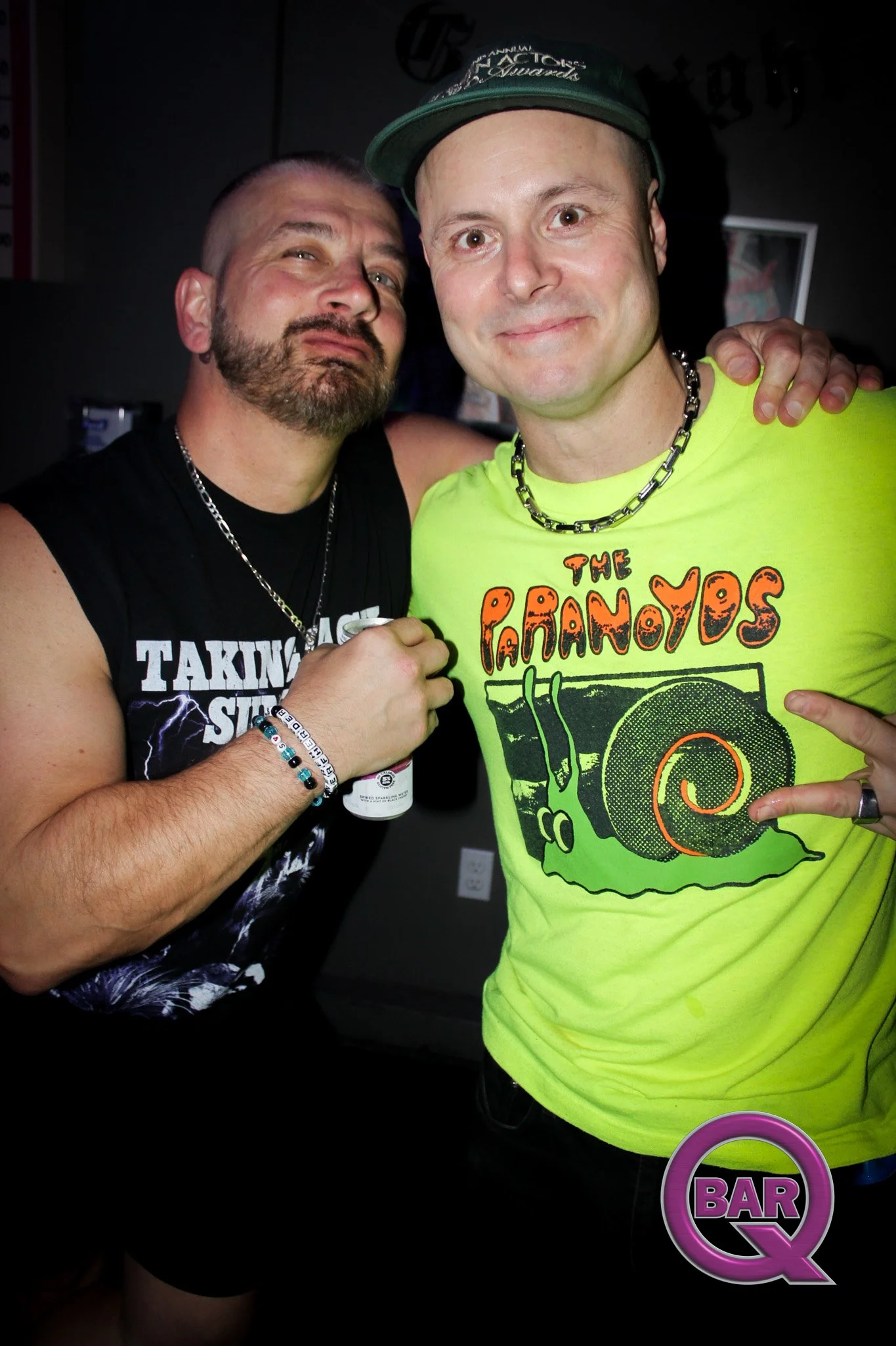 Two men posing for a photo, one wearing a black sleeveless shirt and the other a bright yellow shirt with a graphic design, both smiling and showing hand gestures. The man in yellow is wearing a necklace and a cap. There is a neon bar sign that reads