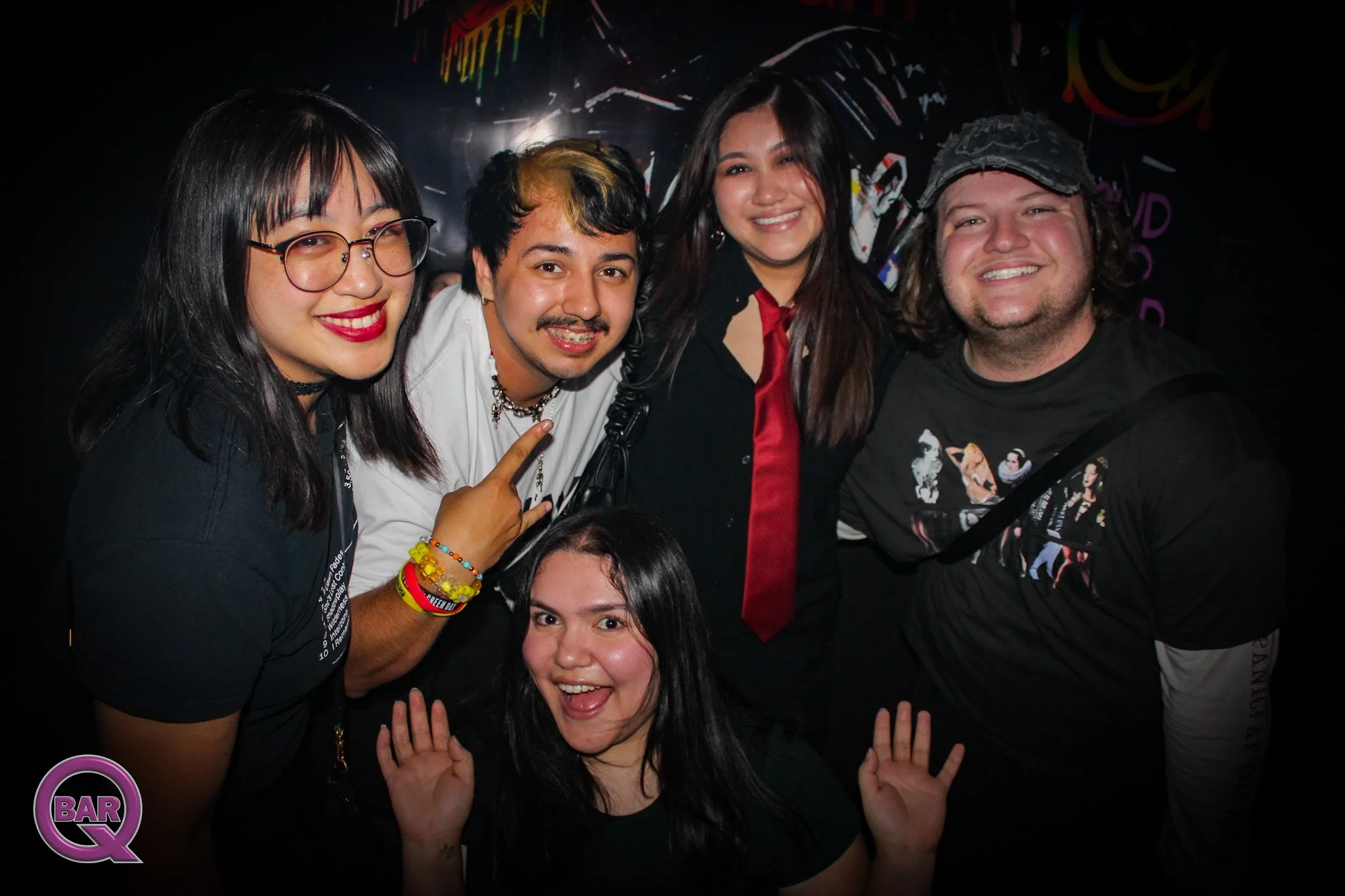 Group of five diverse people smiling and posing together at a bar, with a dark background and colorful lights.