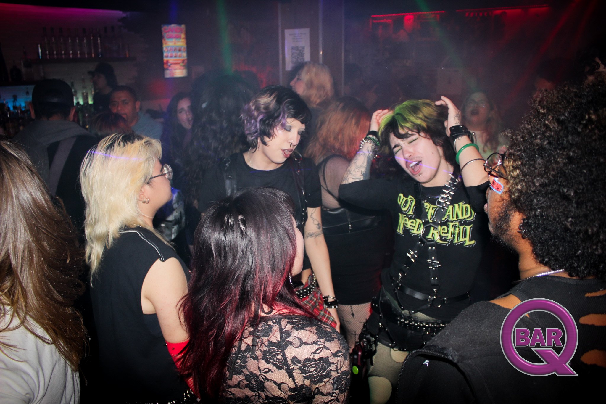 A group of people dancing and socializing in a dimly lit bar or nightclub with colorful lighting and a neon sign in the background.