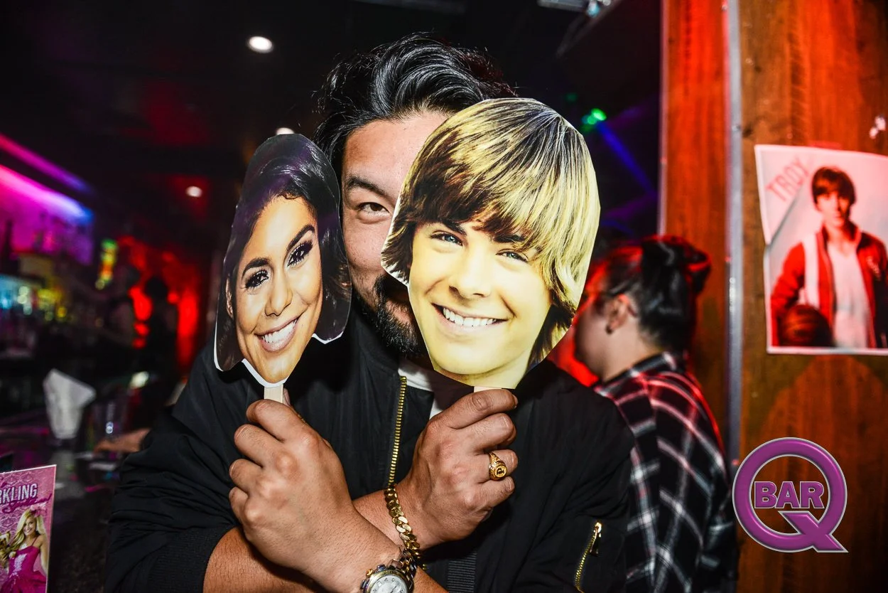 Person holding cutouts of two faces, a woman and a young man, in a bar or nightclub with colorful lighting and other people in the background.