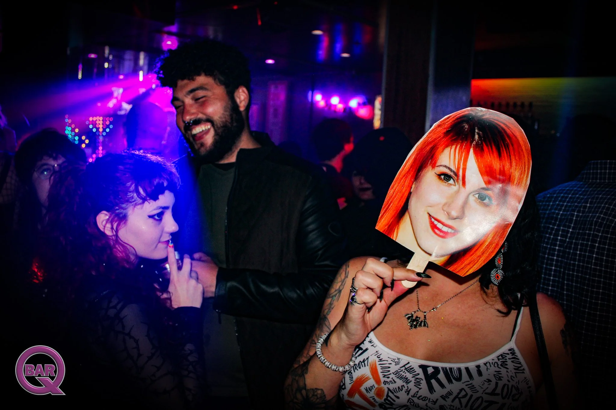People dancing and socializing in a nightclub with colorful lights. A woman with orange hair is holding a mask of a woman with long red hair.