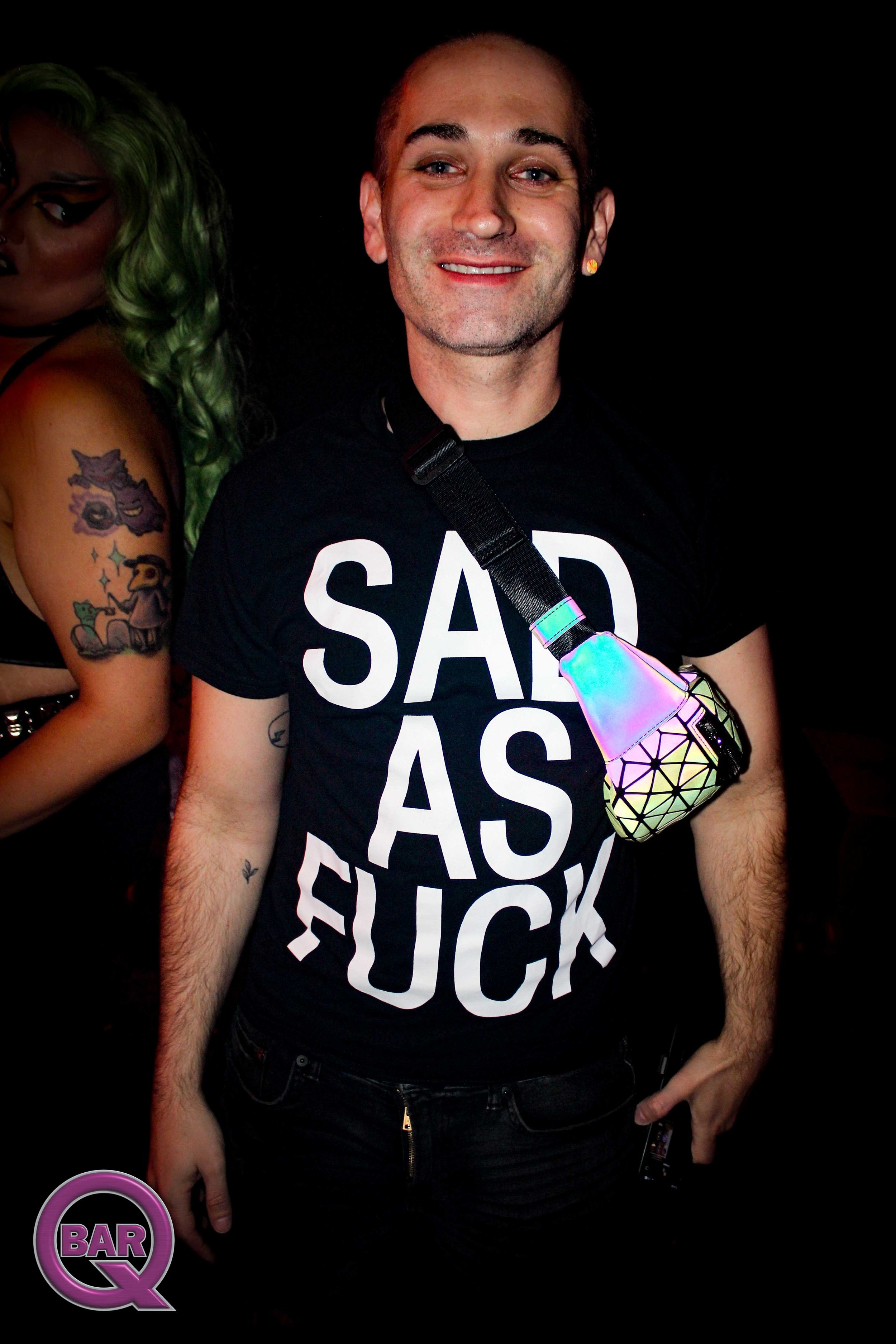 A smiling man wearing a black T-shirt with large white text, a rainbow-colored holographic crossbody bag, and an earring. Partially visible woman with green hair and tattoos is next to him, in a dark party setting.
