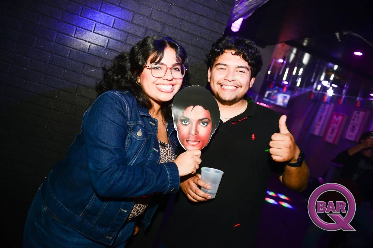 Two smiling friends at a bar, holding a Michael Jackson photo mask, with neon lights and a black brick wall in the background.