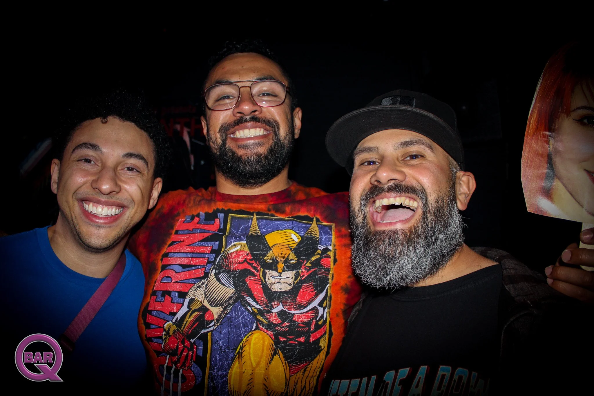 Four men smiling and posing together in a dark setting, with one holding a phone and a poster partially visible.
