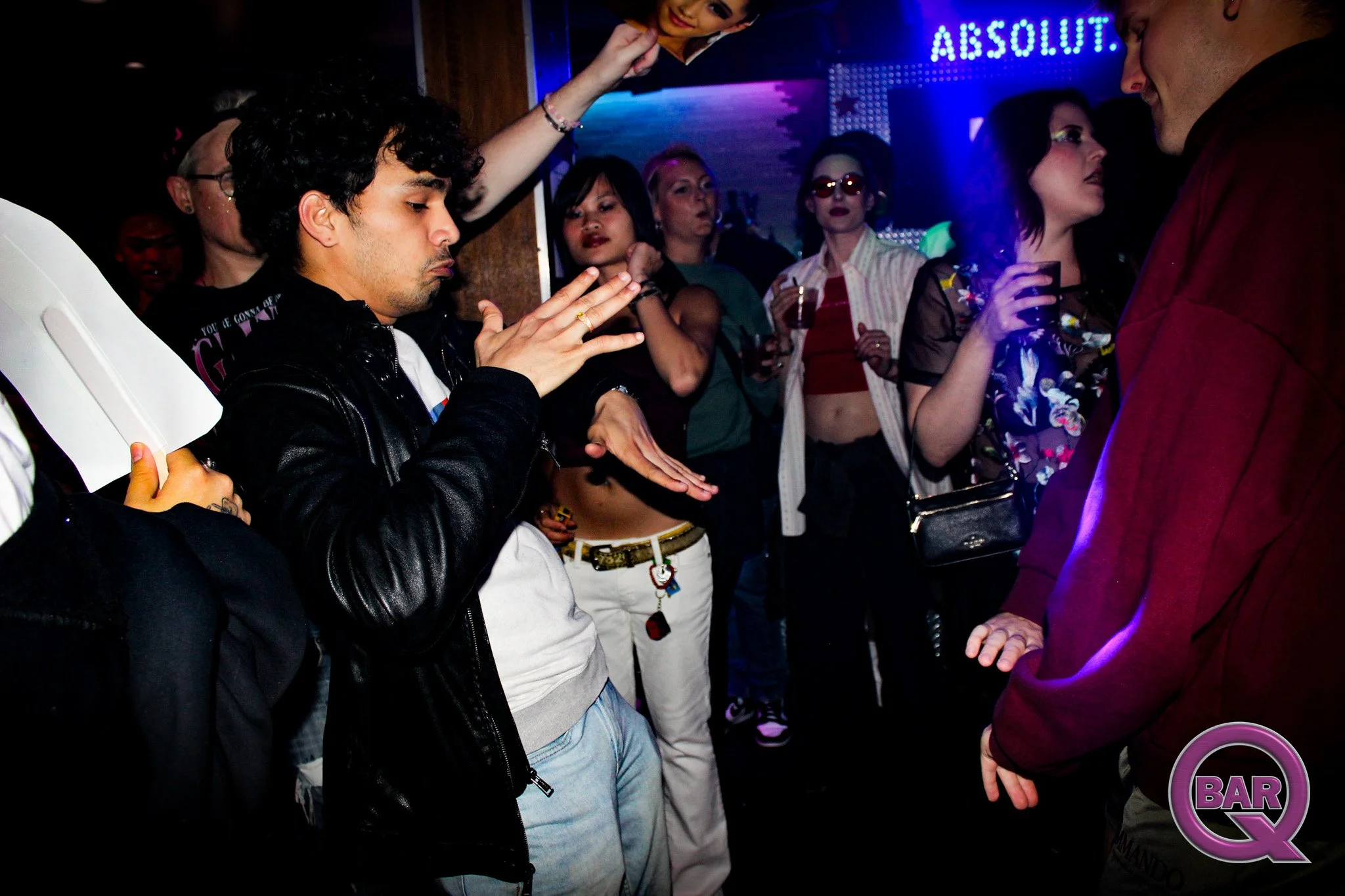 A group of people at a bar or nightclub, some with drinks, standing close together in a dark, lively setting with colorful lighting and a neon sign in the background.