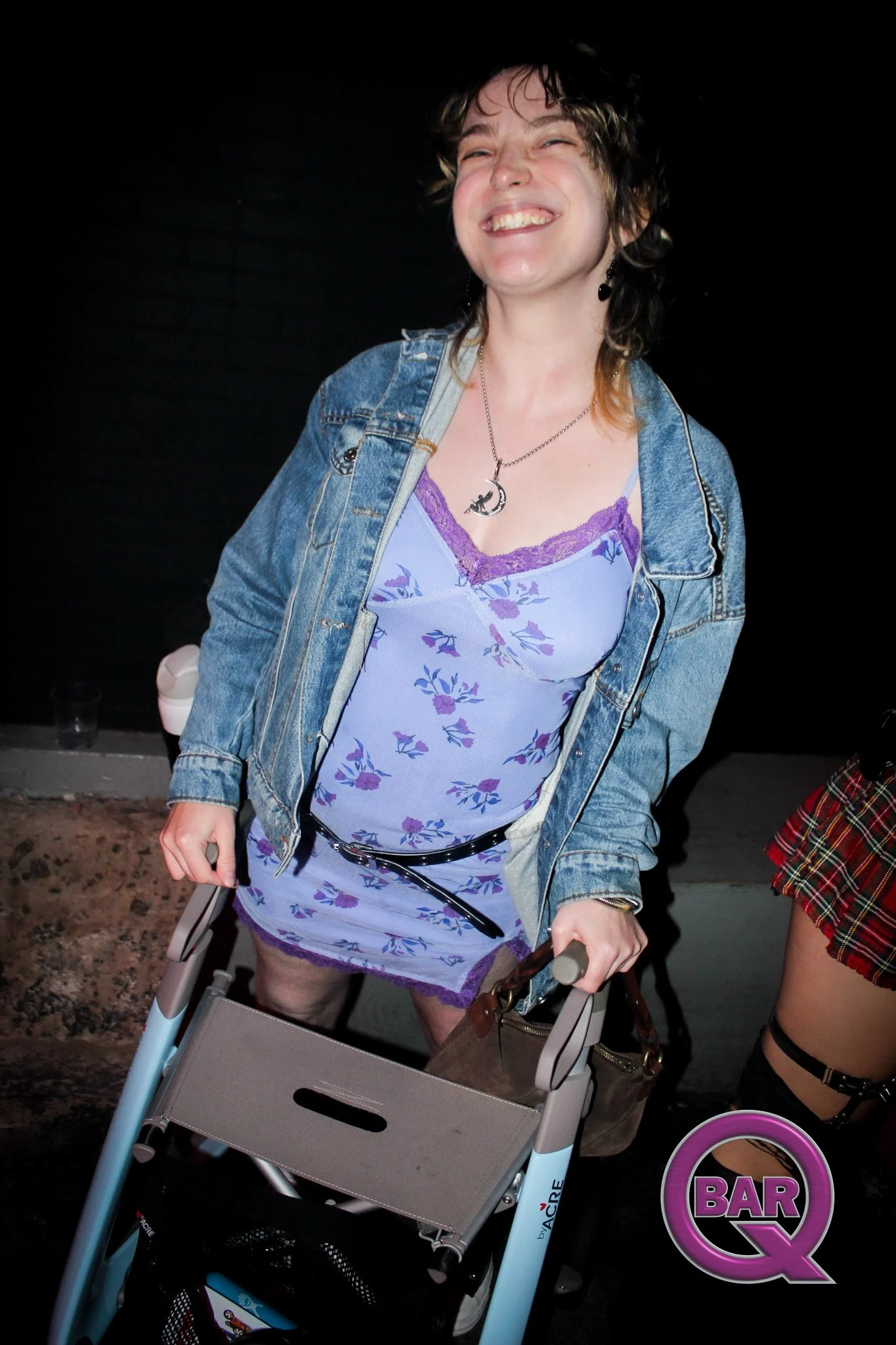 Woman smiling wearing a purple floral dress, denim jacket, and moon necklace, pushing a gray stroller at nighttime.
